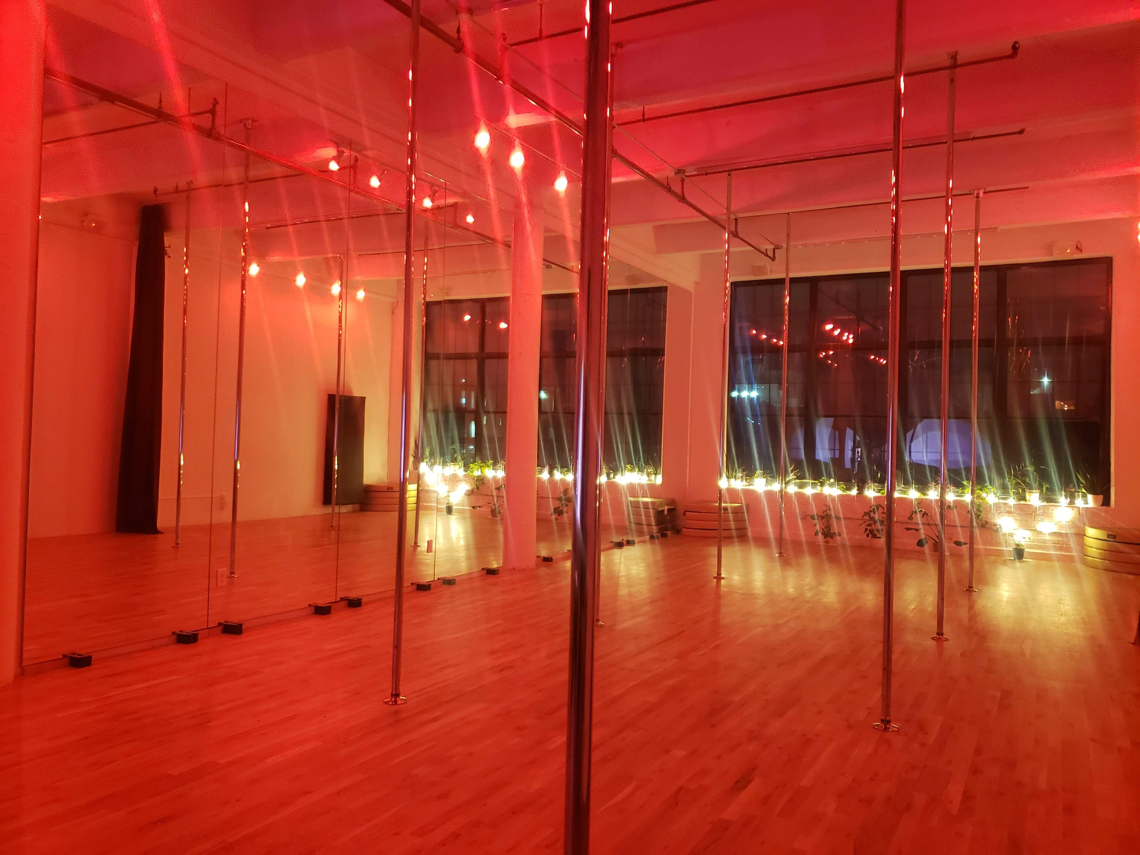 A dance studio with multiple polished poles and red lighting, featuring wooden flooring and large windows illuminated by soft lights.