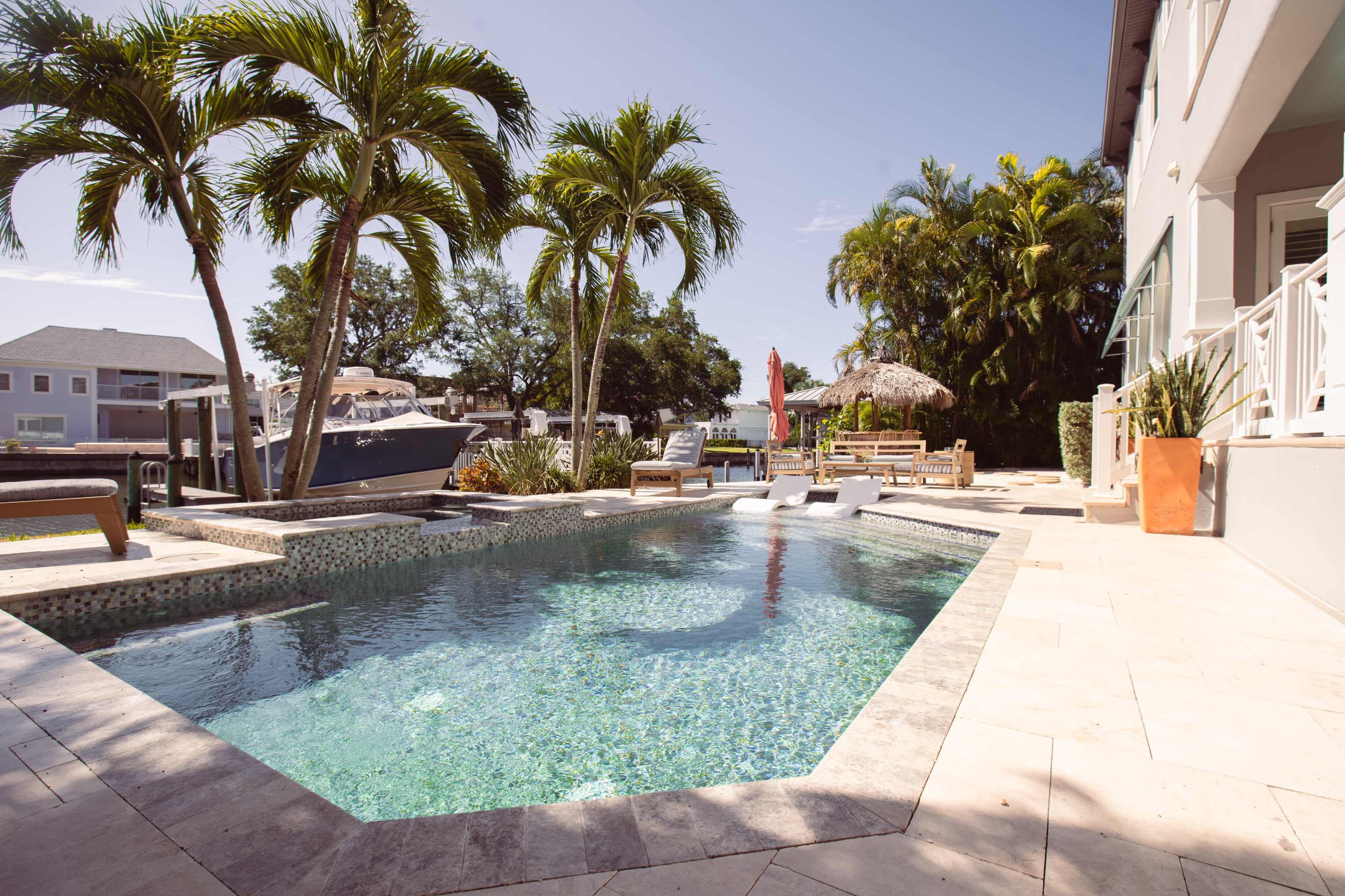 The image showcases a modern outdoor pool surrounded by palm trees and lounge furniture, set against a waterfront view.