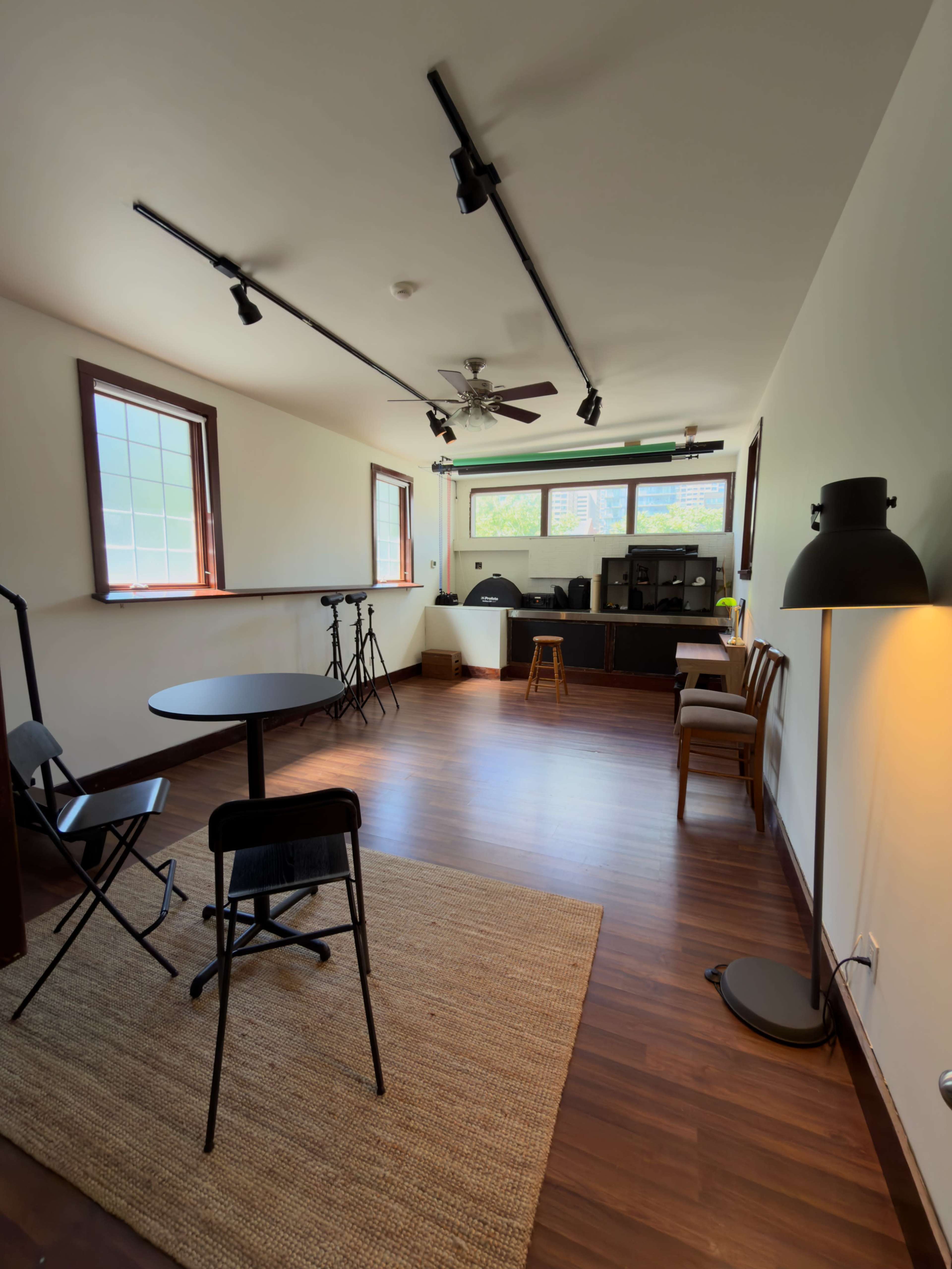 The image shows a spacious room with wooden floors, featuring a round table and chairs, a small stool, and a workspace against one wall with audio equipment and track lighting.