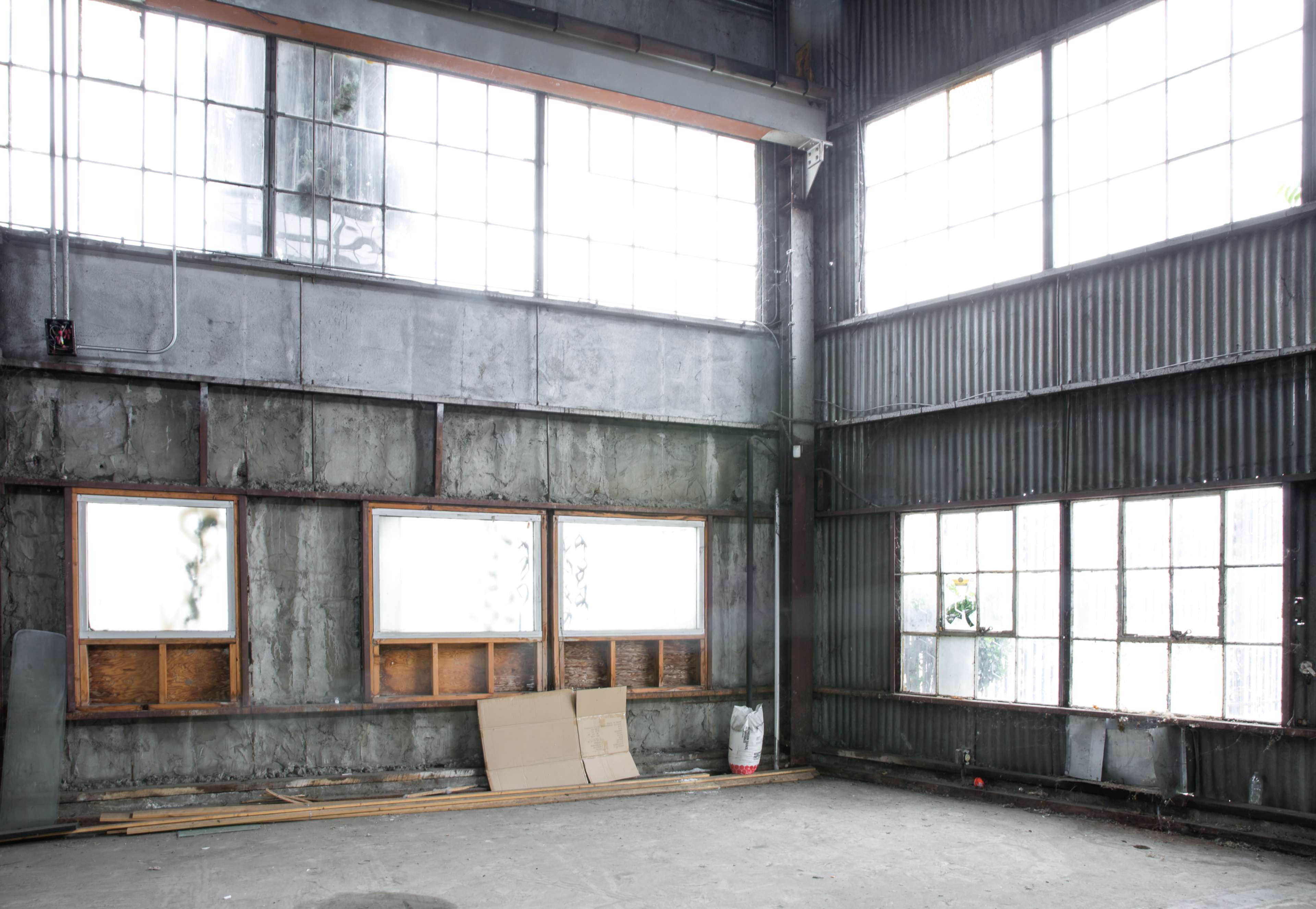 An empty, industrial space with large windows and bare concrete walls, featuring wooden frames and some scattered materials on the floor.