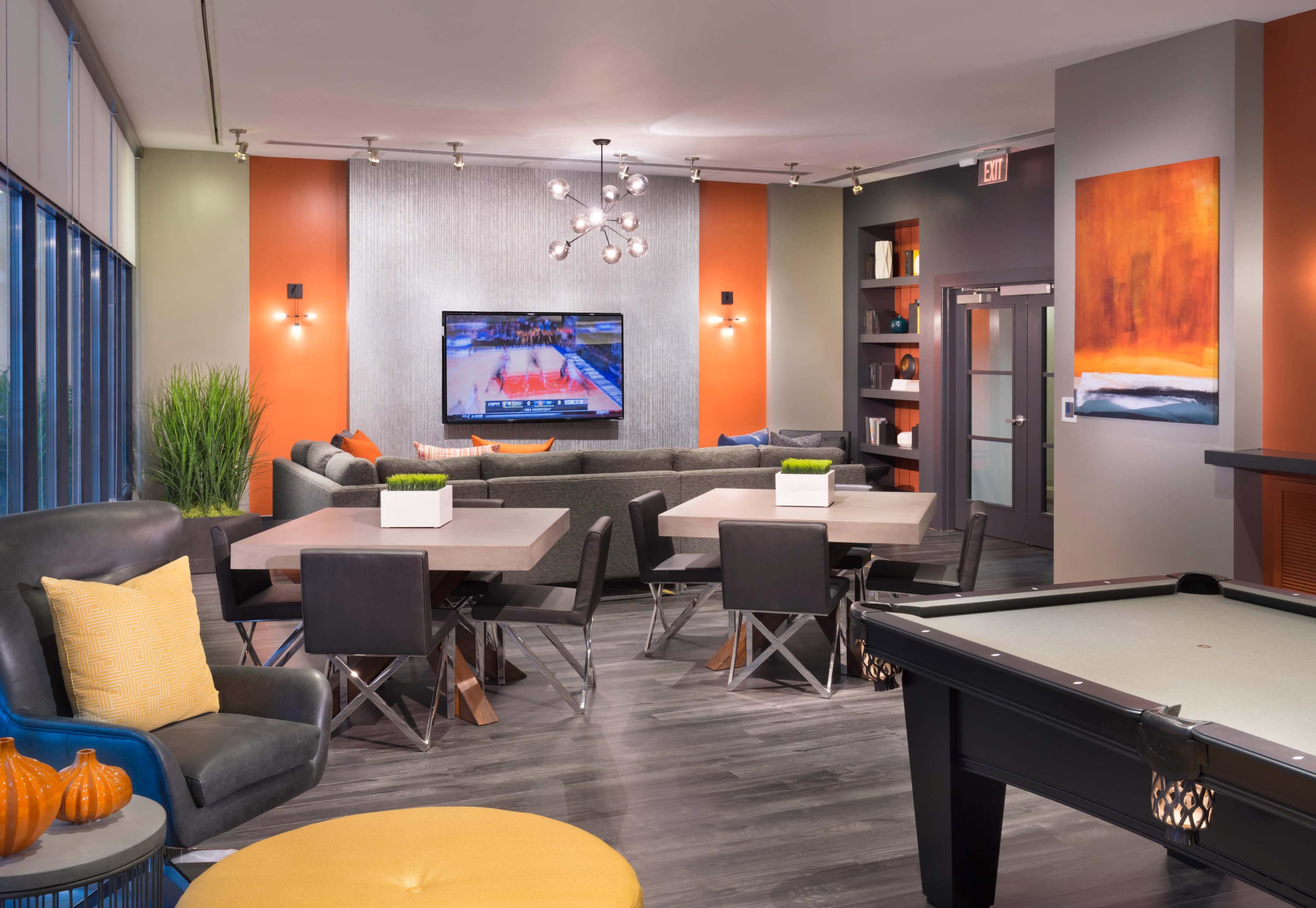 The scene features a modern lounge area with a large sectional sofa, tables, a pool table, and a television displaying a sports game against a backdrop of colorful wall decor.