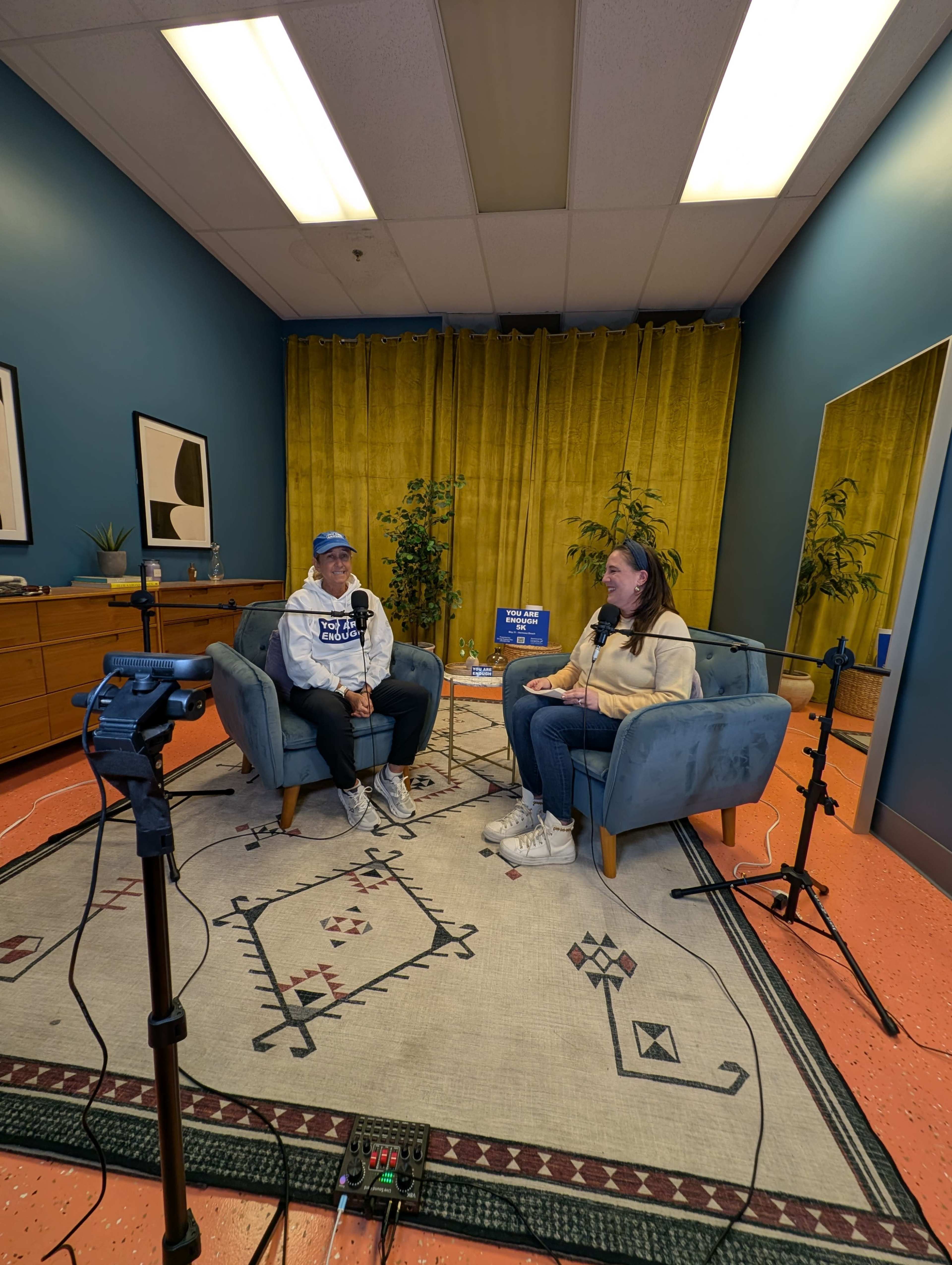 A man and a woman sit in a cozy recording studio with microphones in front of them, surrounded by blue walls and yellow curtains.