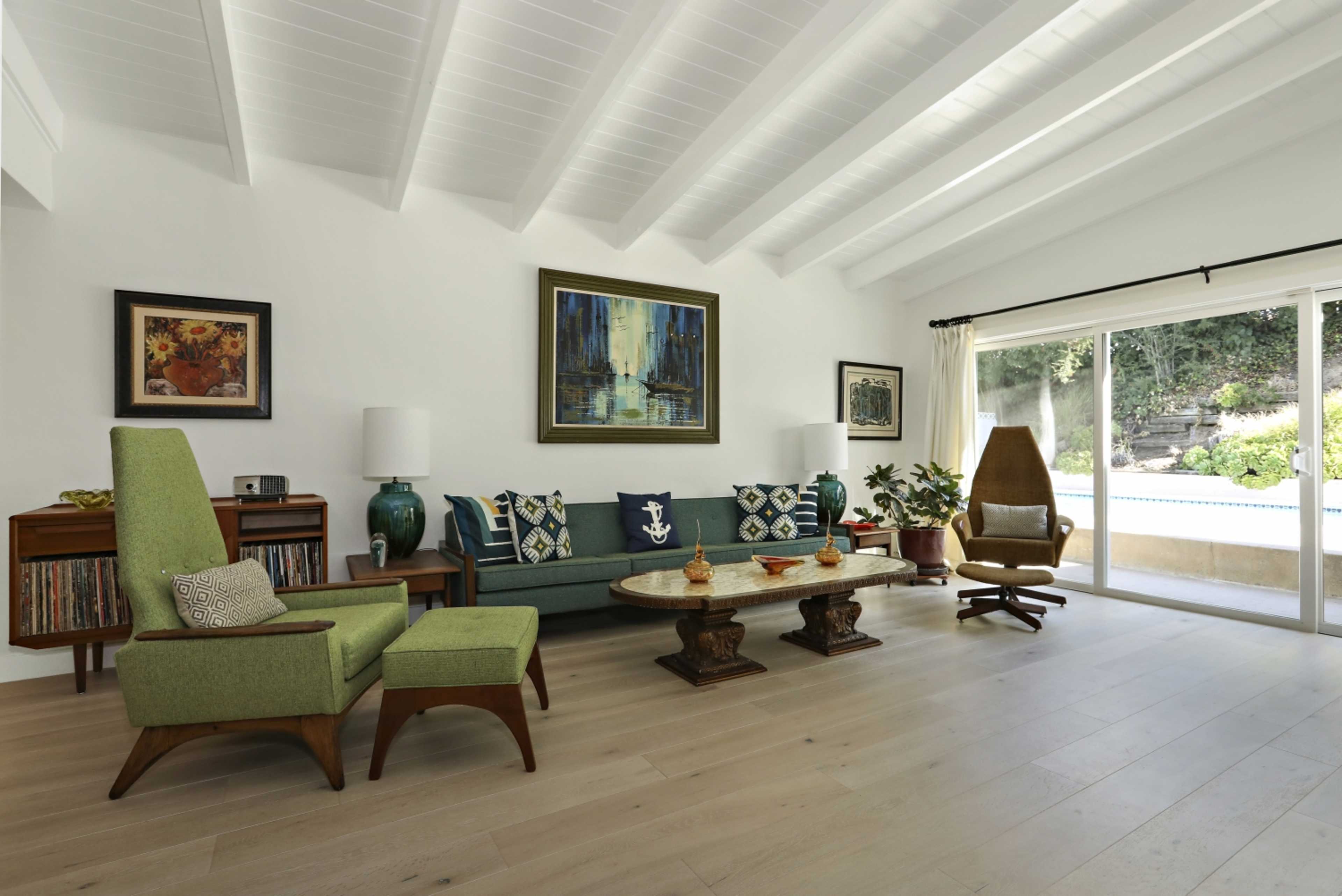 The living room features a green sofa and armchair, a round coffee table, and large windows overlooking an outdoor area.