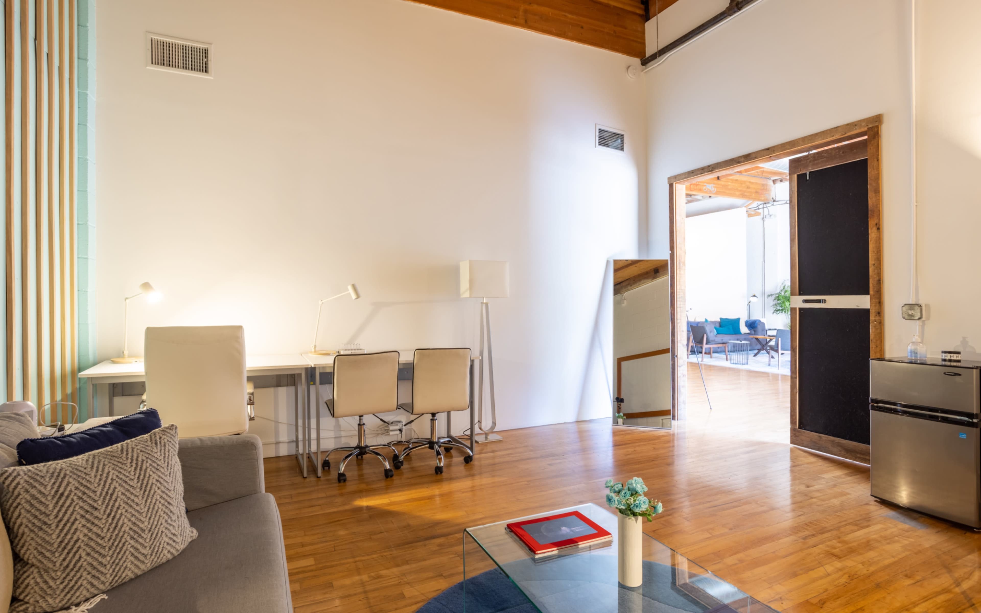 A modern, open-concept living space features a seating area, a desk with chairs, a large mirror, and a doorway leading to another room.
