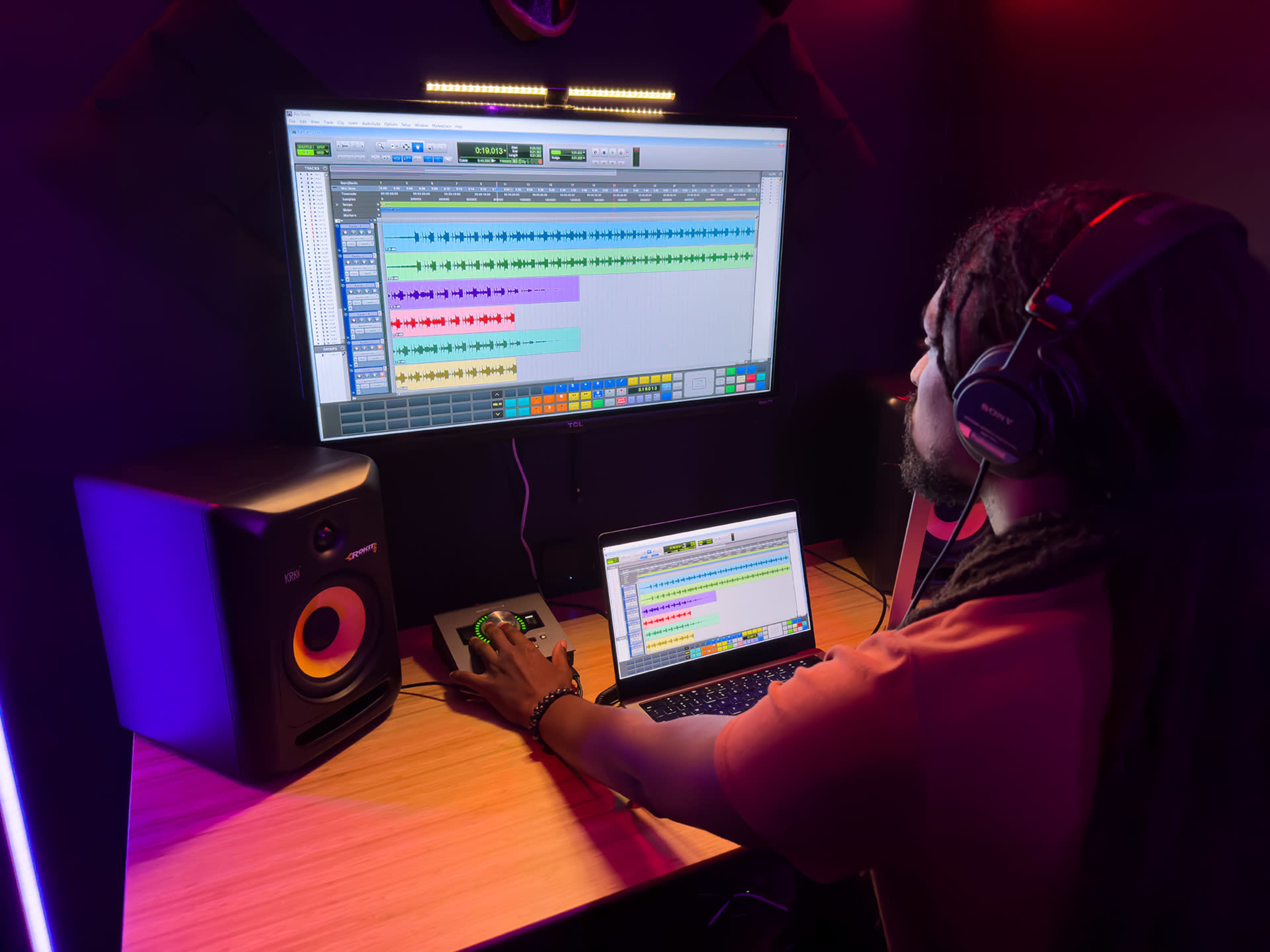 A person is working at a desk with a laptop and a large monitor displaying music production software, while speakers are positioned on either side.