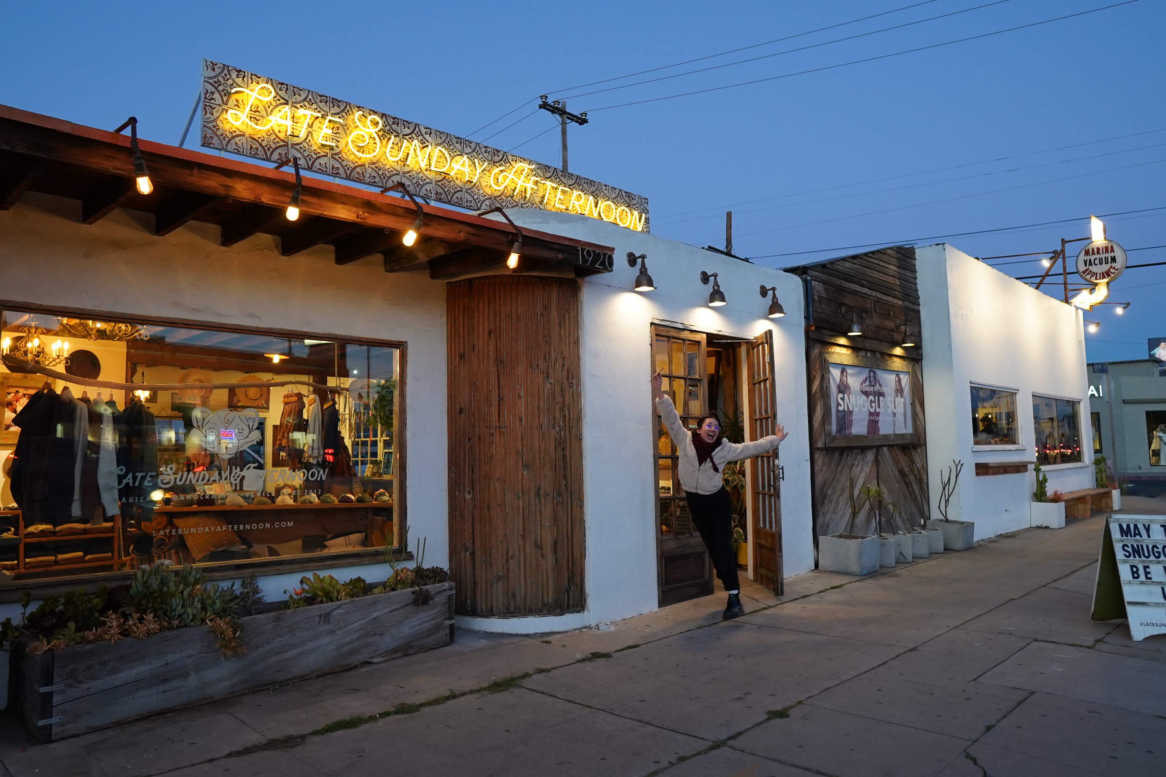 A person stands outside a stylish retail shop with a sign that reads "Late Sunday Afternoon" at dusk, showcasing its welcoming entrance and large windows.