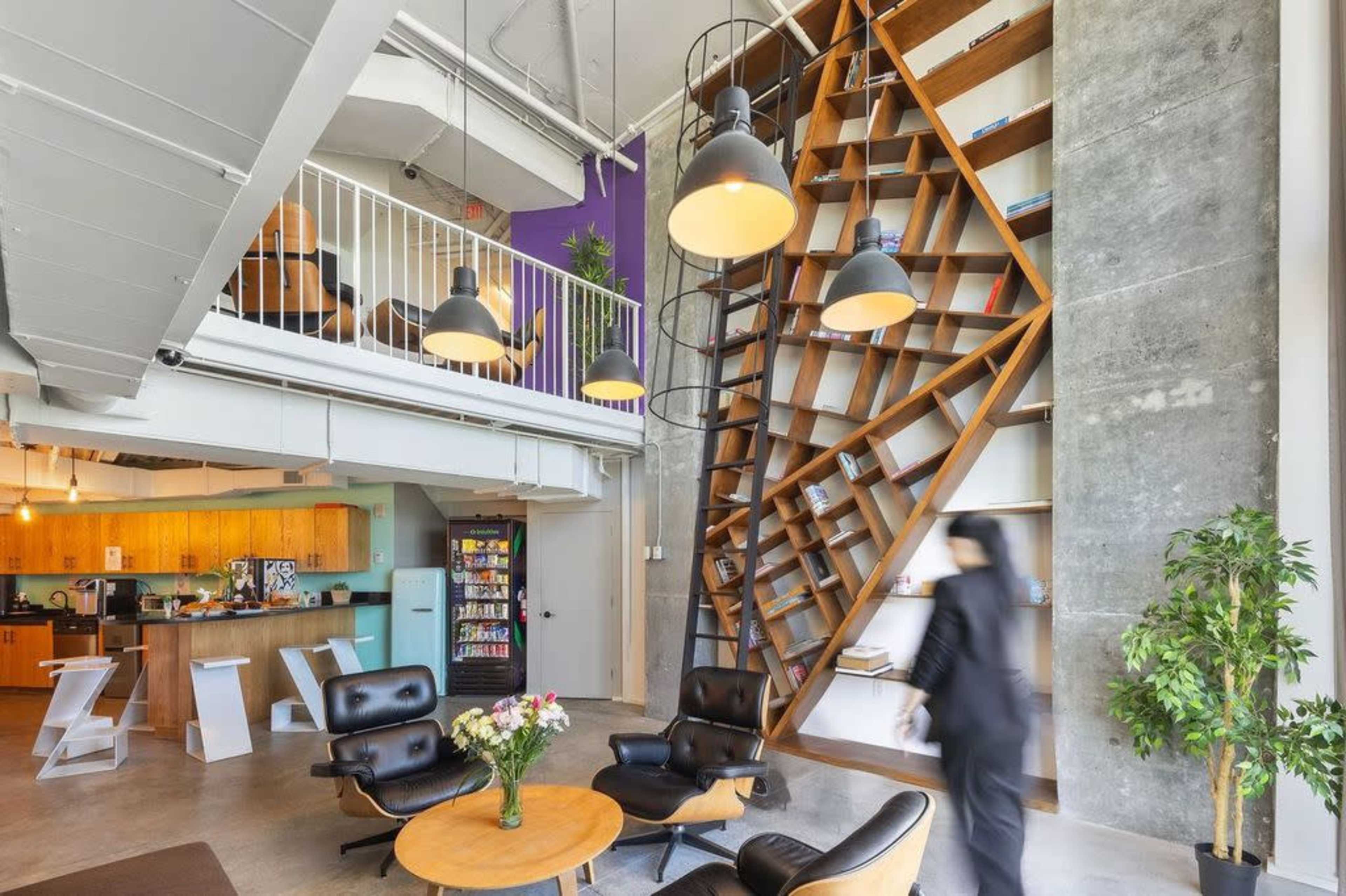 A modern open-concept office space with a spiral bookshelf, a kitchen area, and a seating arrangement featuring black chairs and a round table.