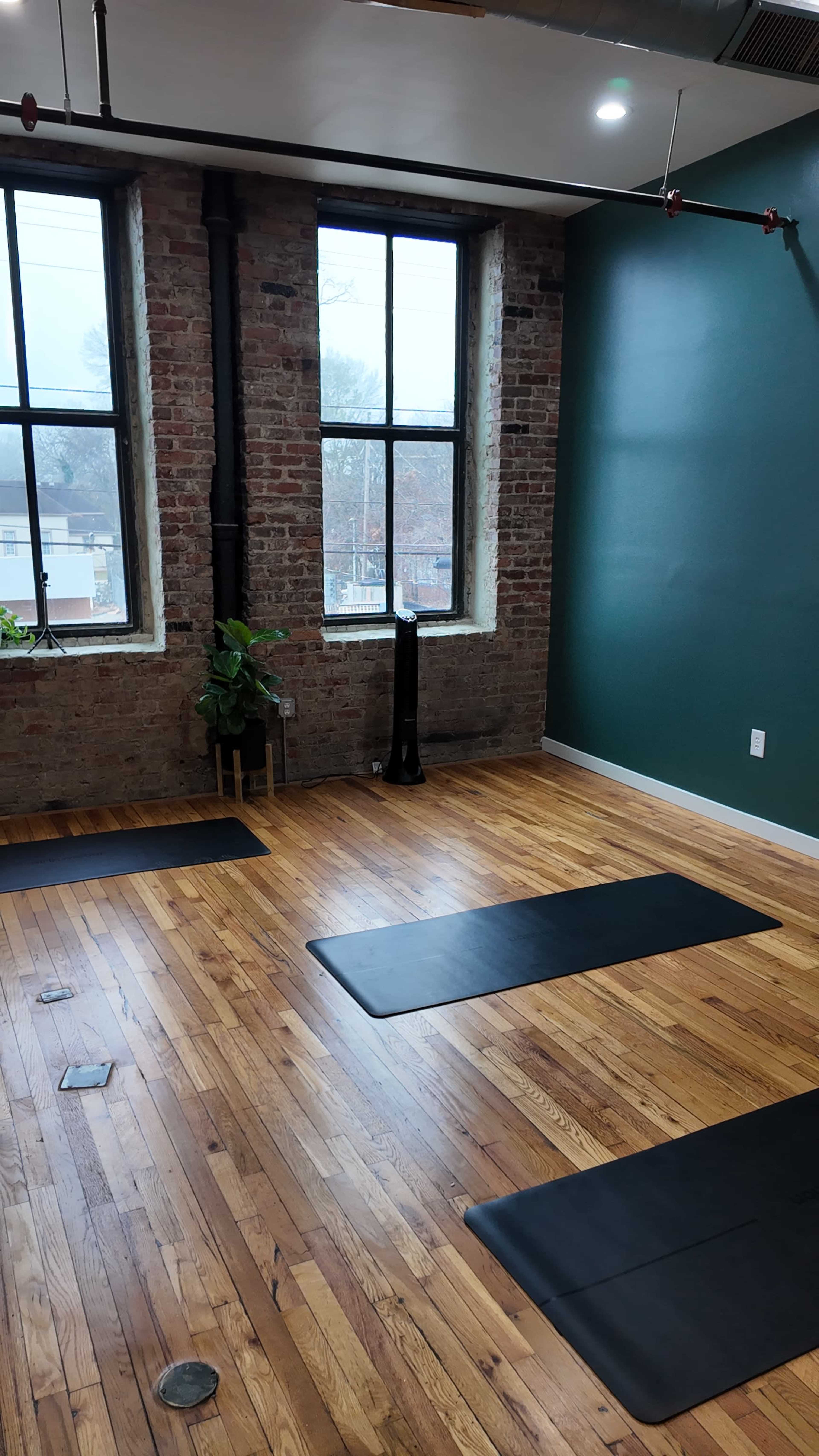 The image shows an open room with wooden floors, large windows, and yoga mats placed on the floor.