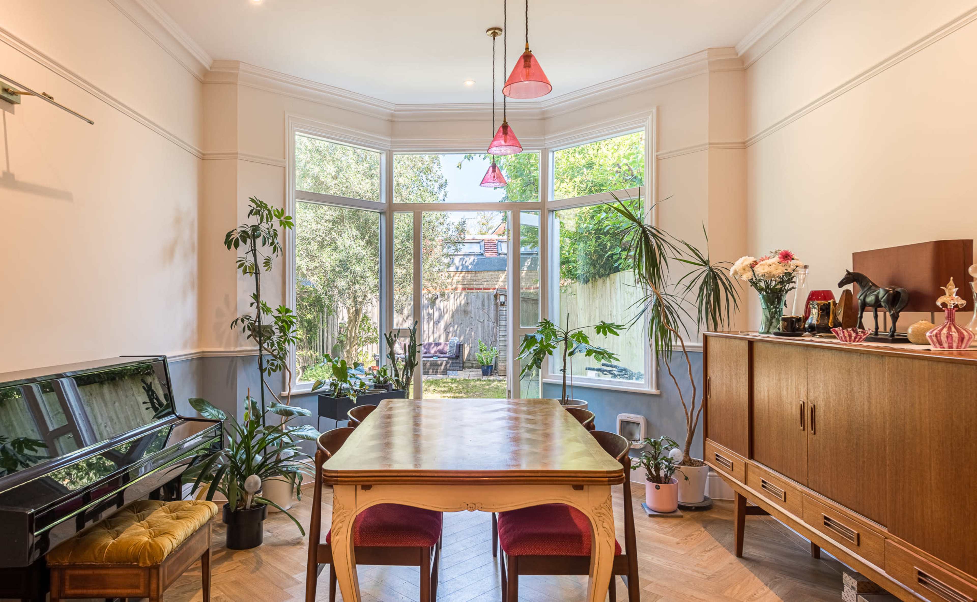 The image shows a bright dining area with a wooden table, chairs, and plants, adjacent to large windows overlooking a garden.