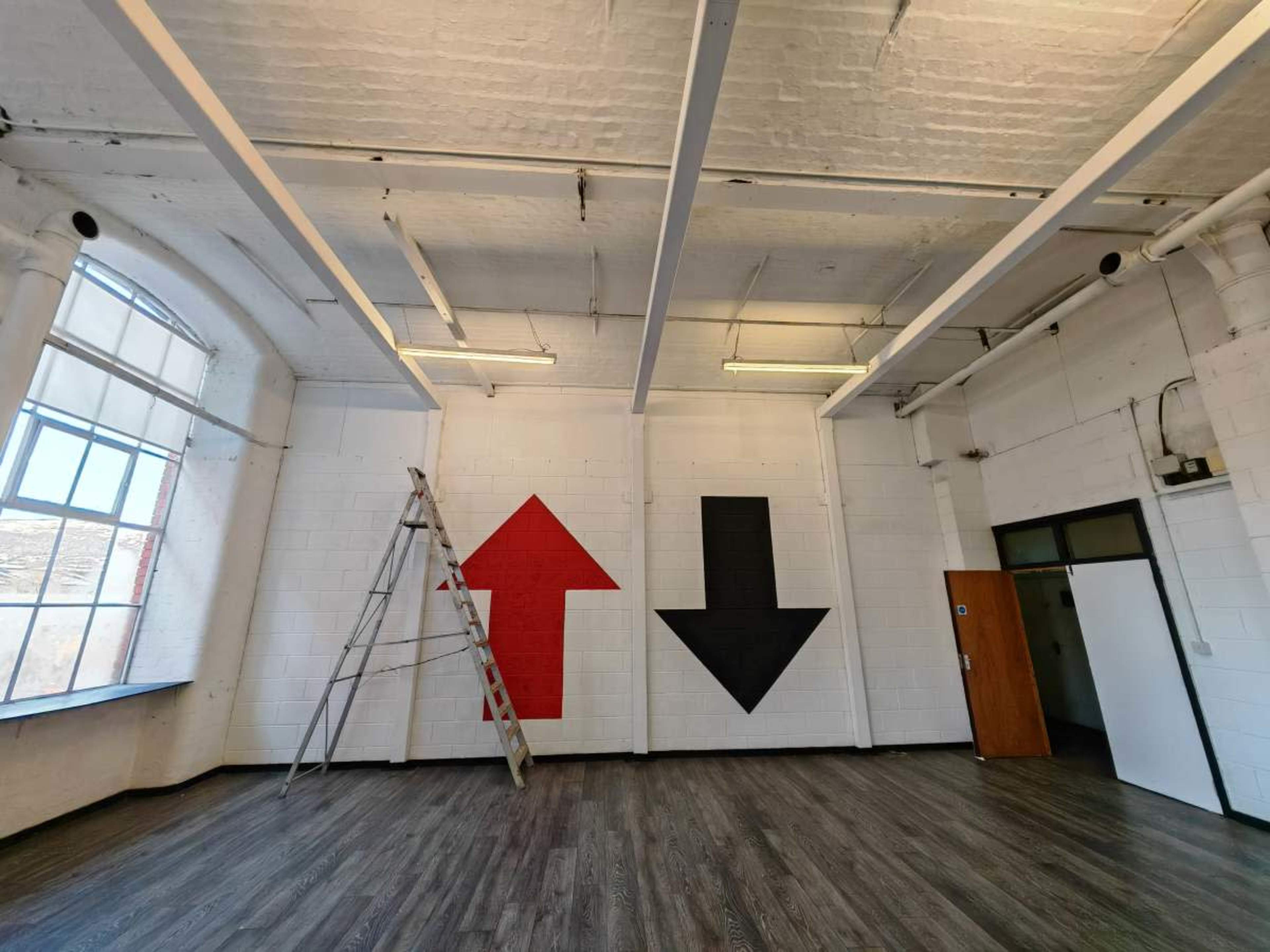 The image shows an empty room with wooden flooring, featuring two large arrows painted on the walls—one red pointing up and one black pointing down—alongside a ladder leaning against the wall.