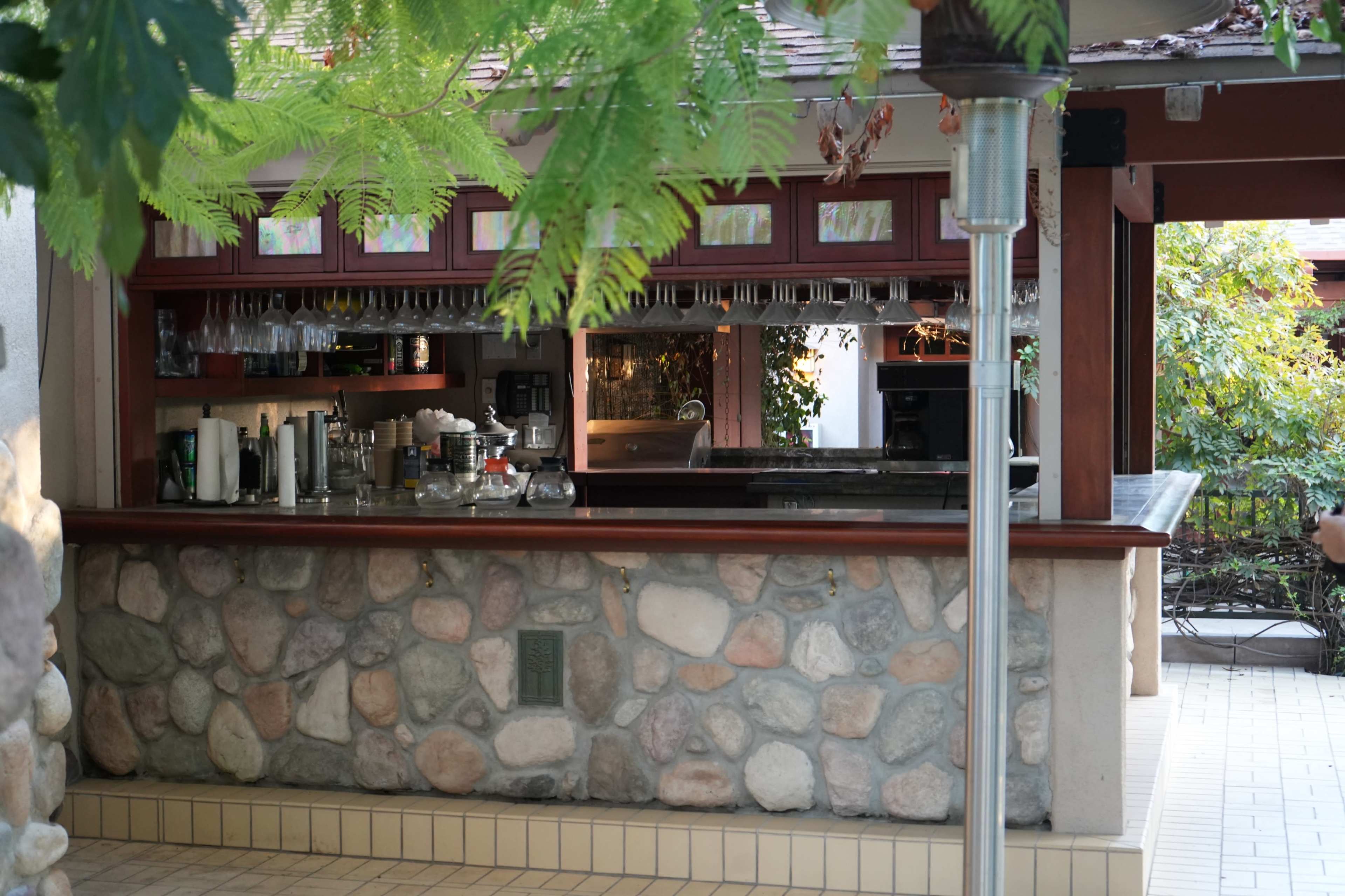 The image shows an outdoor bar with a stone counter, behind which are shelves stocked with glasses and drink-making equipment.