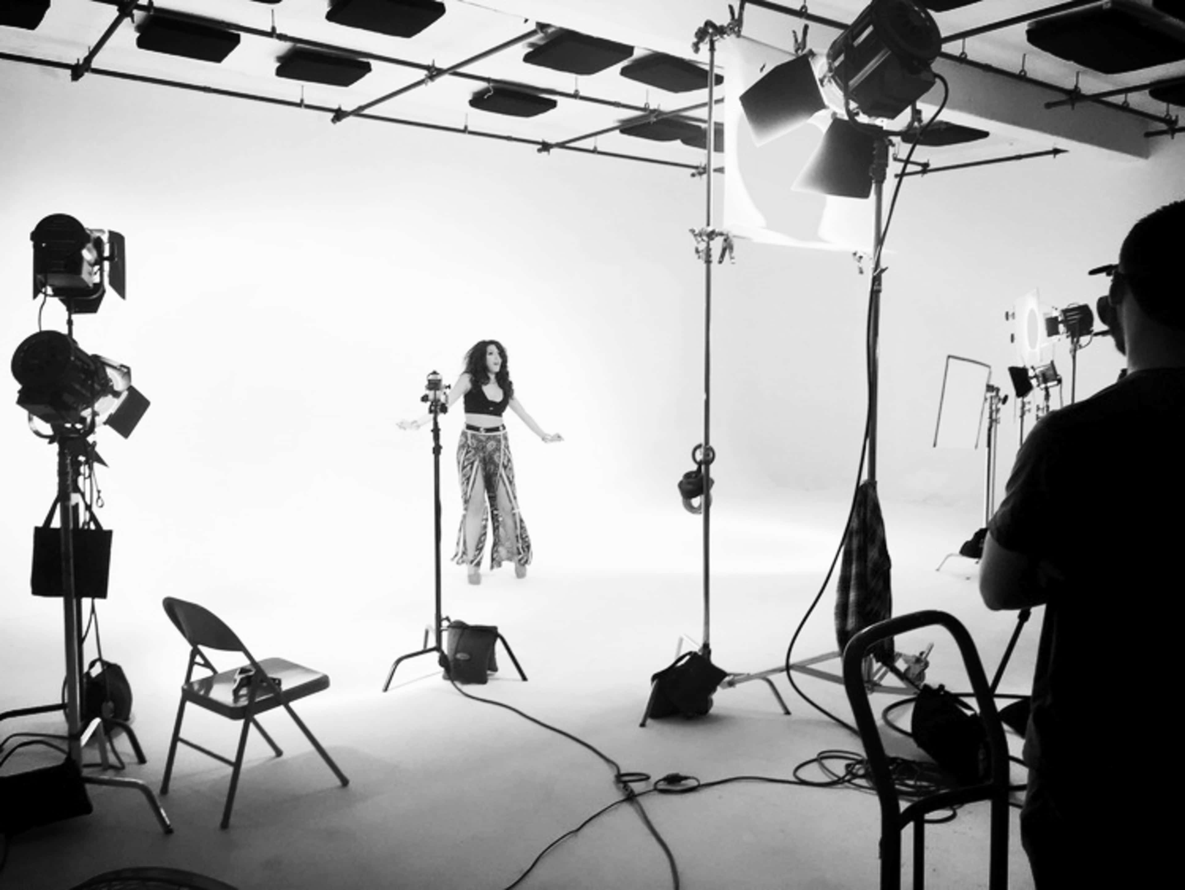 A woman wearing a black crop top and patterned pants stands in the center of a photography studio, surrounded by bright lights and camera equipment.