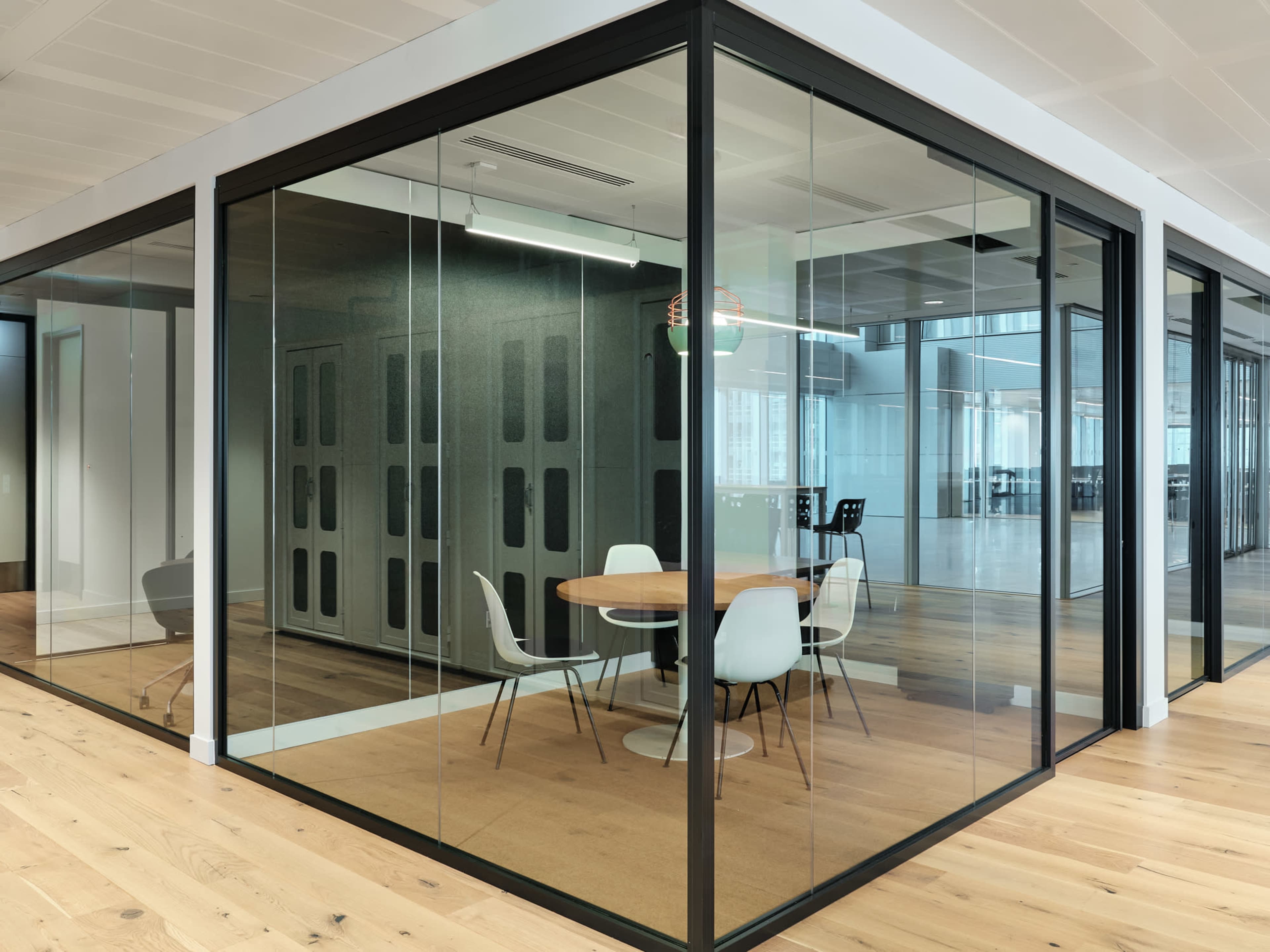 A glass-walled meeting room features a circular table surrounded by four chairs on a wooden floor.