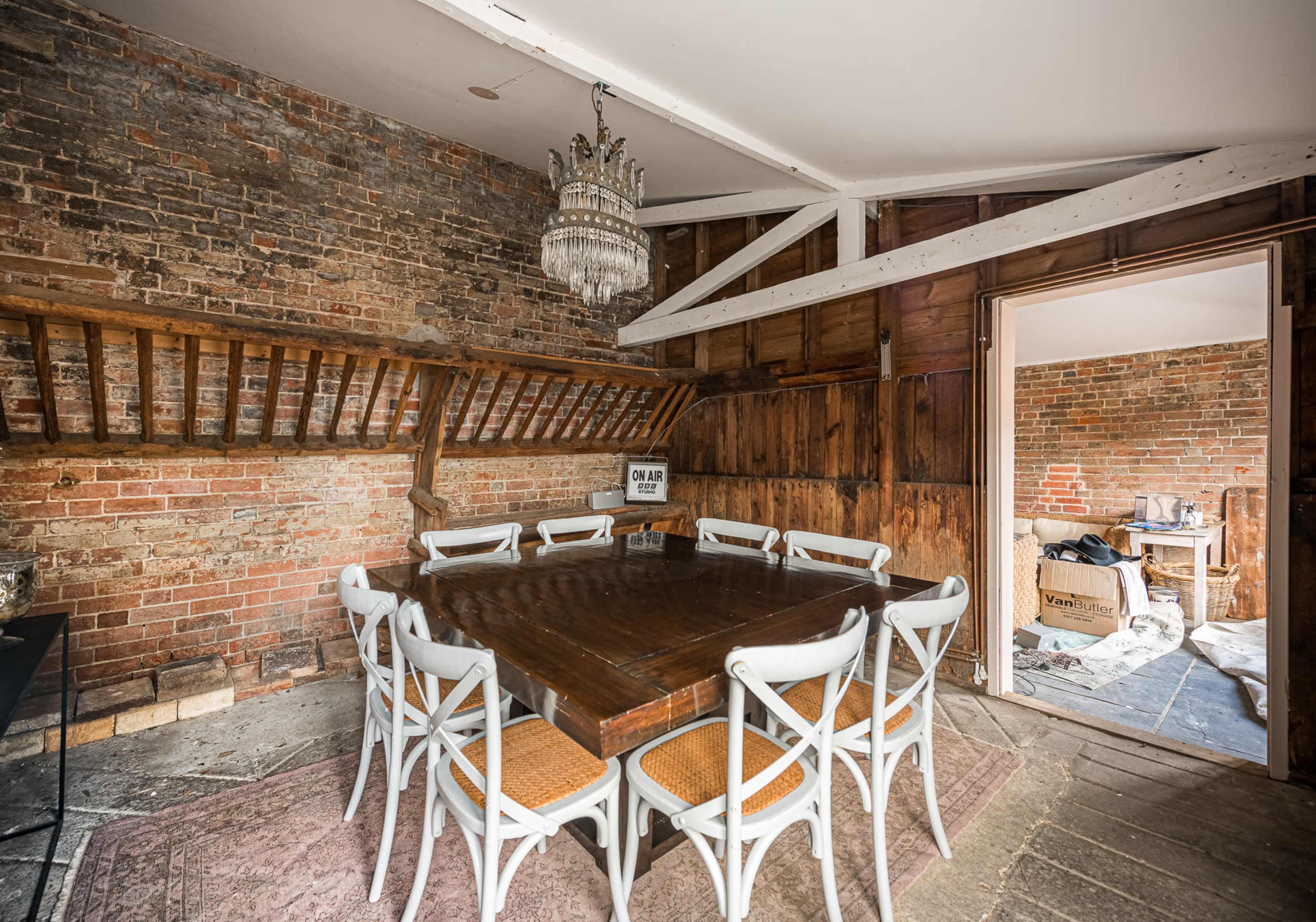 The image shows a rustic dining room featuring a large wooden table surrounded by twelve white chairs, with exposed brick walls and a chandelier overhead.
