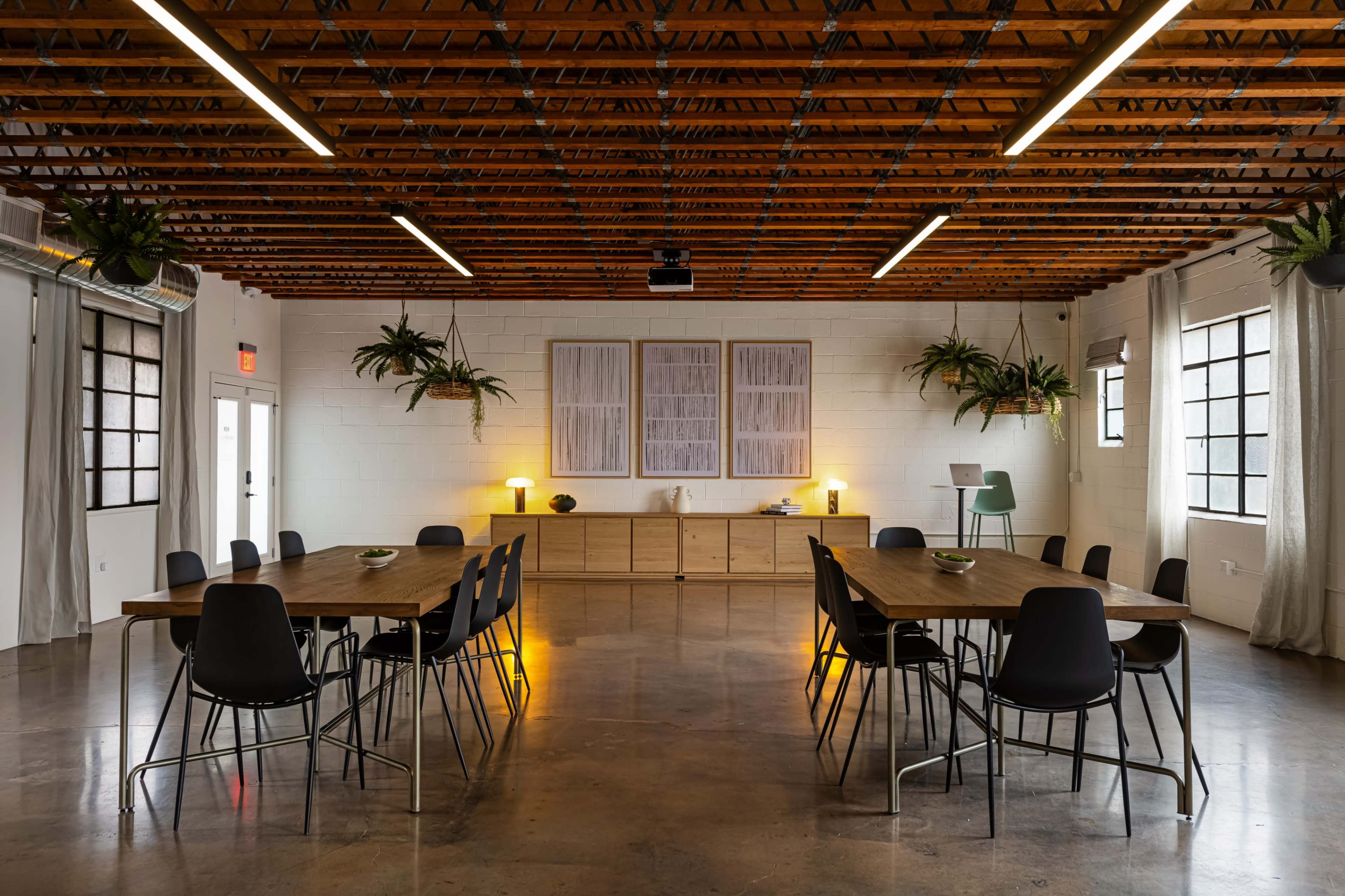 The image shows a spacious meeting room featuring a central round table surrounded by black chairs, with wooden beams overhead and plants hanging from the ceiling.