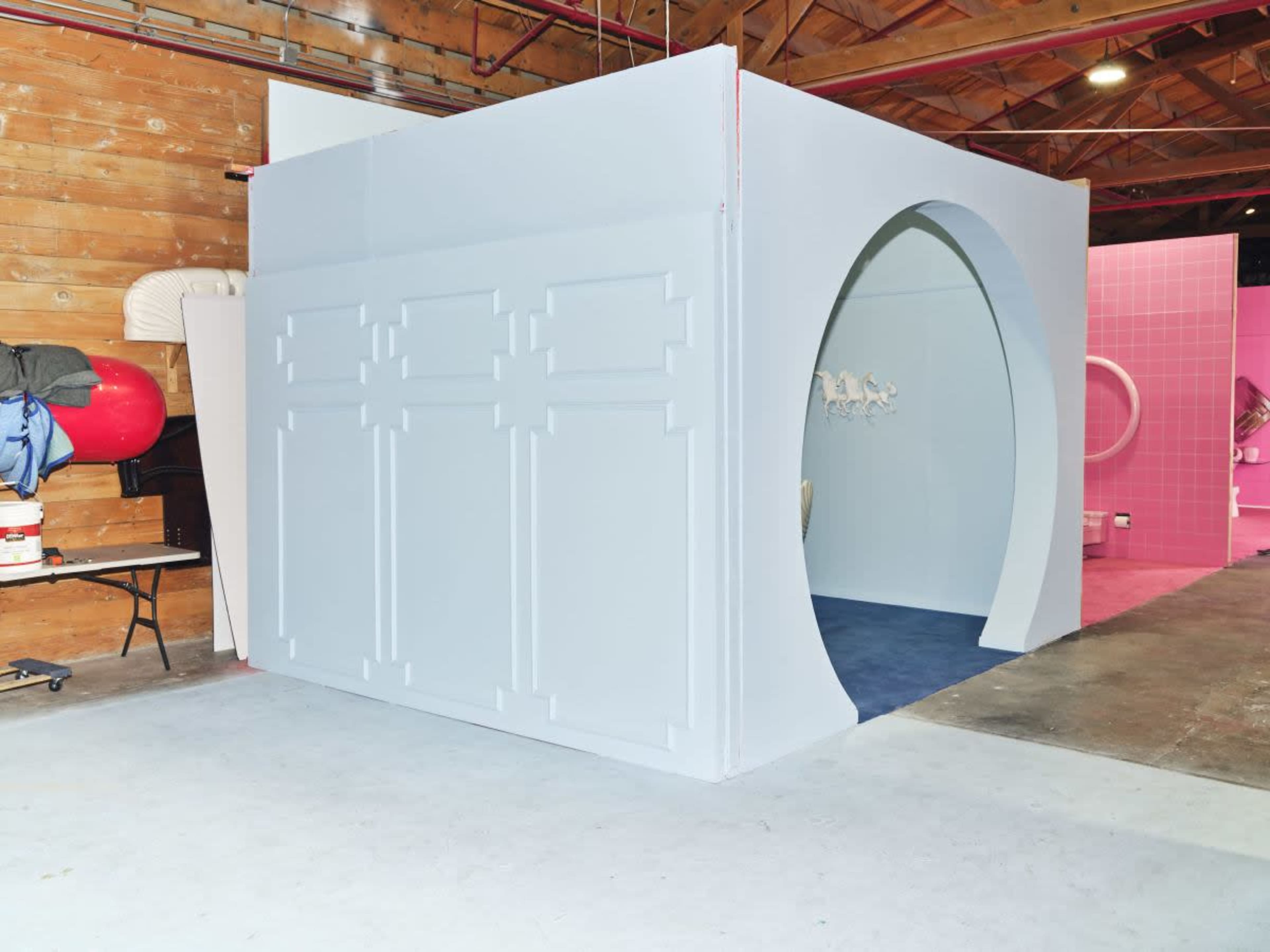 A light blue room partition with a circular entrance, set in an industrial space with wooden walls and concrete flooring.