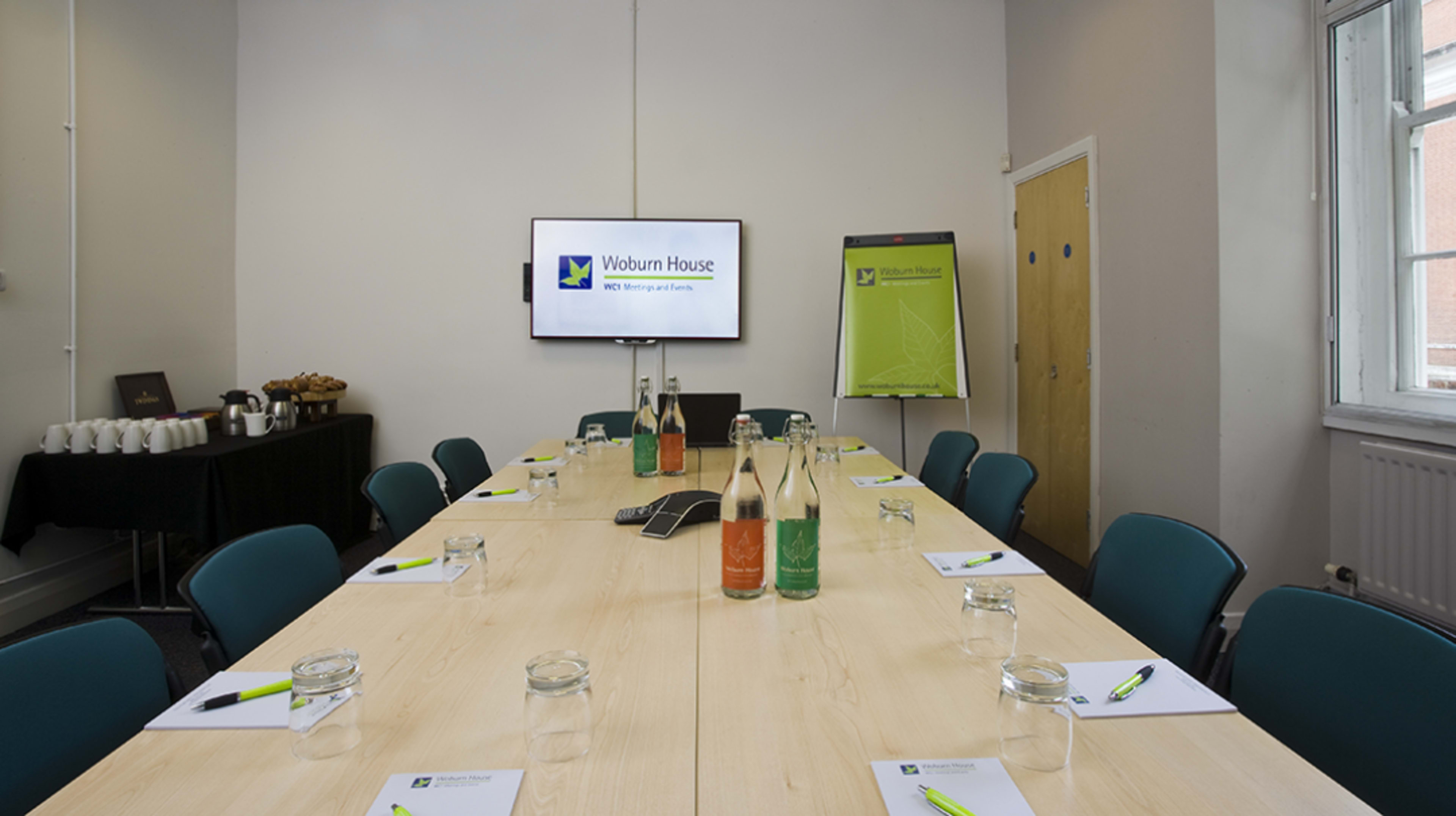 The image shows a long wooden conference table set with notebooks and water bottles, arranged in a meeting room featuring a large screen and a flip chart.