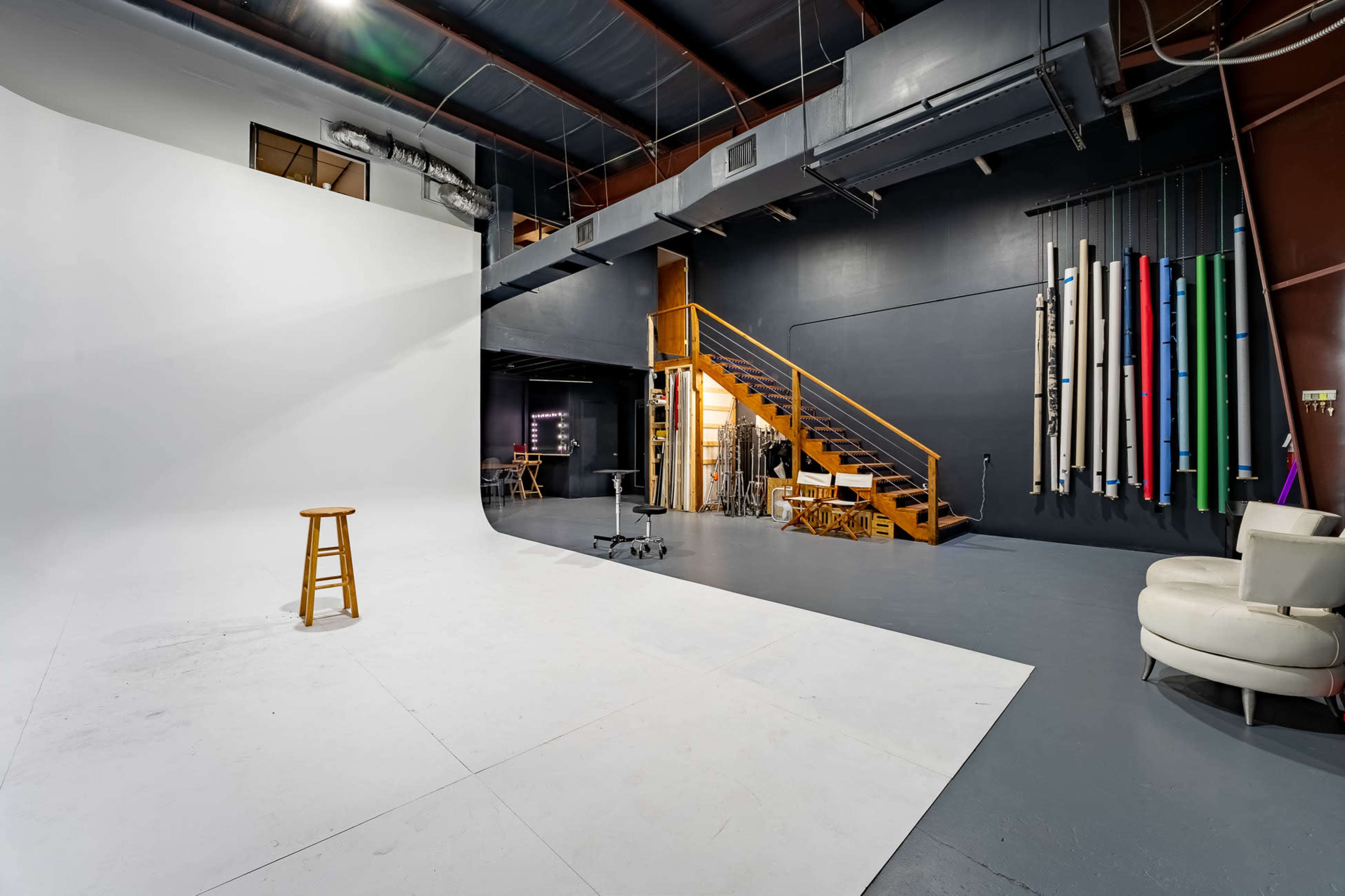 A photography studio with a large white backdrop, a wooden stool, a staircase leading to an upper level, and colorful rolls of backdrop material hanging on the wall.