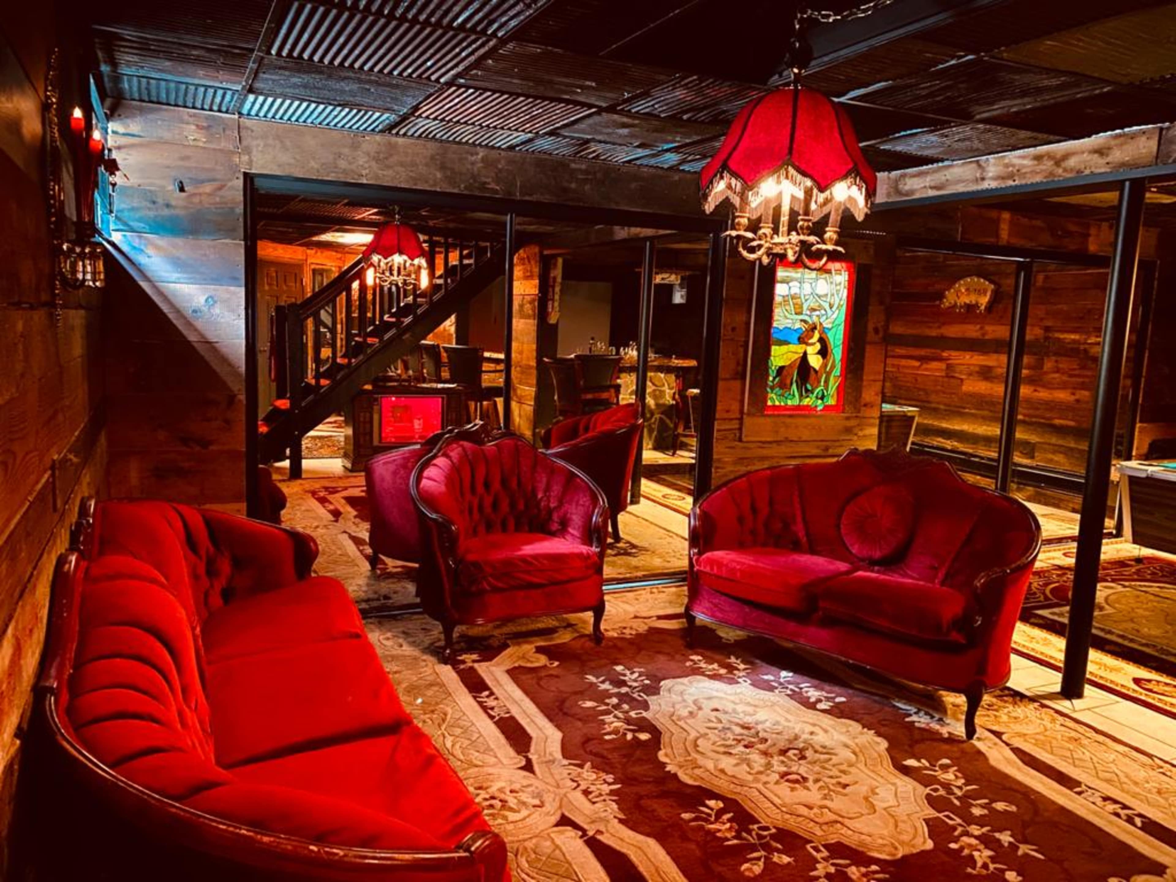 A rustic lounge area featuring red velvet furniture, patterned rugs, and a stained glass window, all illuminated by vintage lamps.