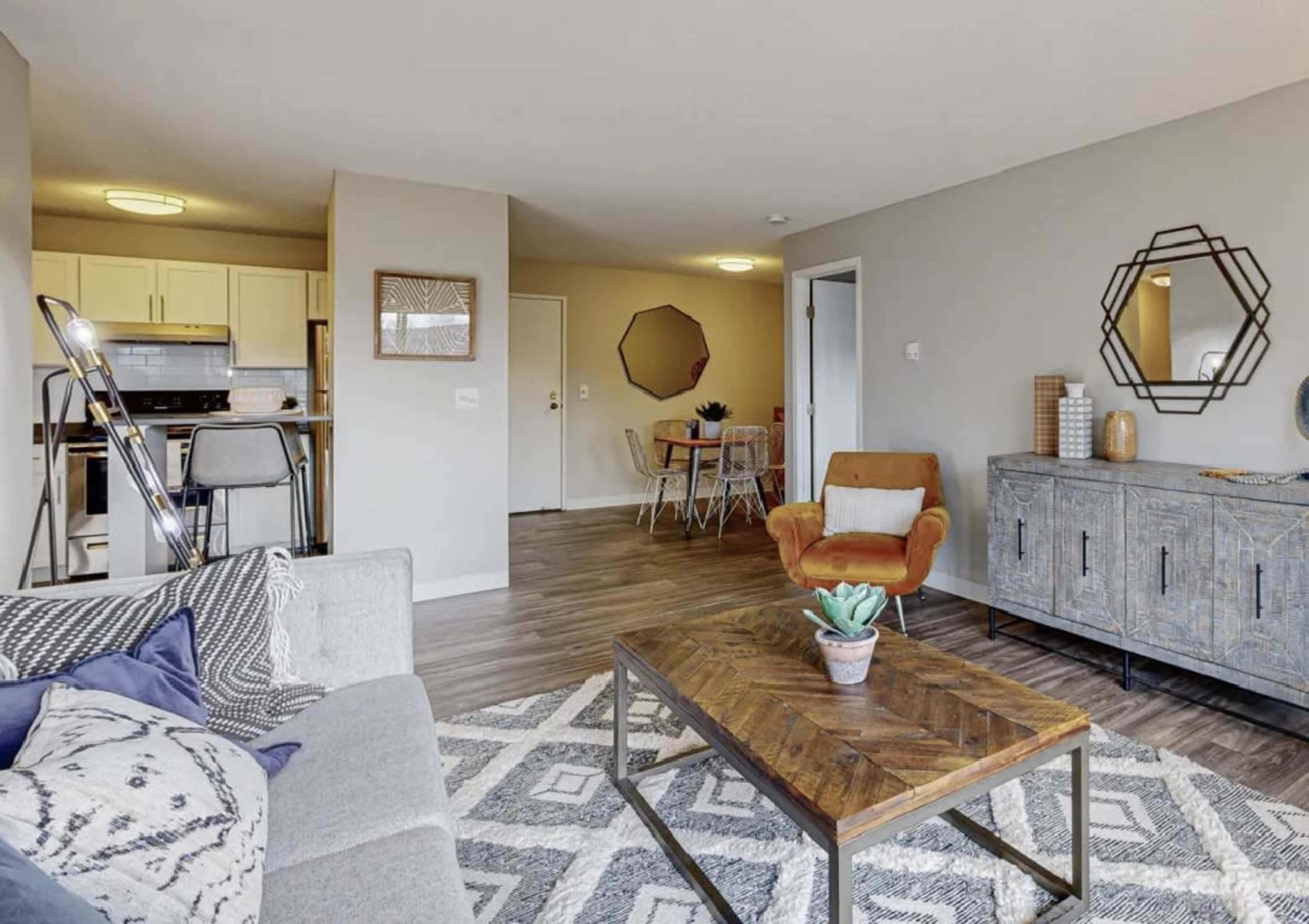 A modern living space featuring a grey sofa, a wooden coffee table with a plant, and a dining area in the background with a round table and chairs.