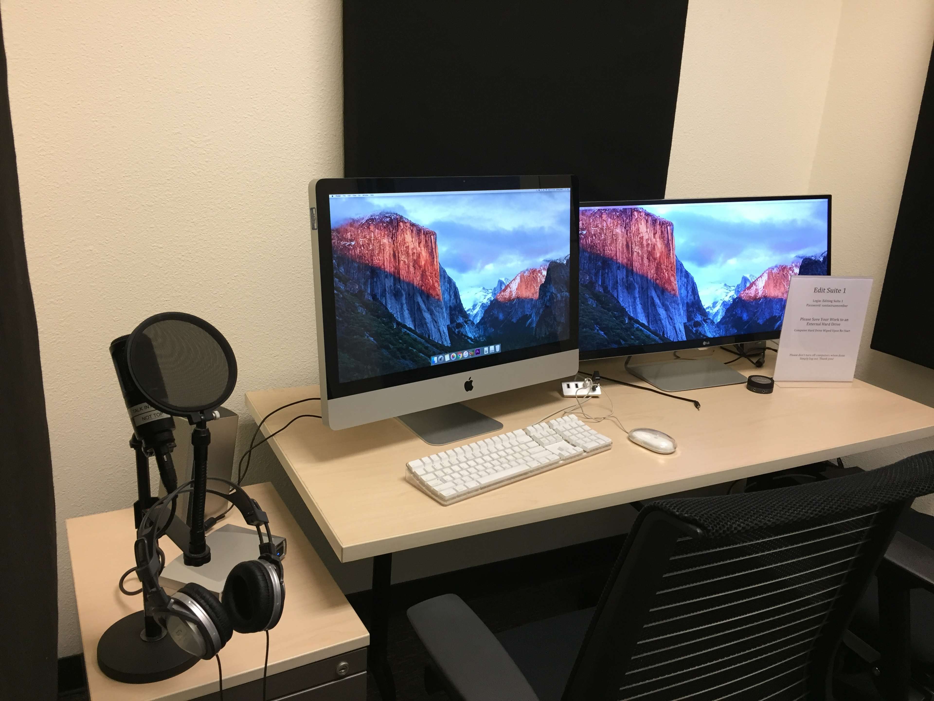 A desk with two computer monitors, a keyboard, headphones, and a microphone, set against a backdrop of black panels.