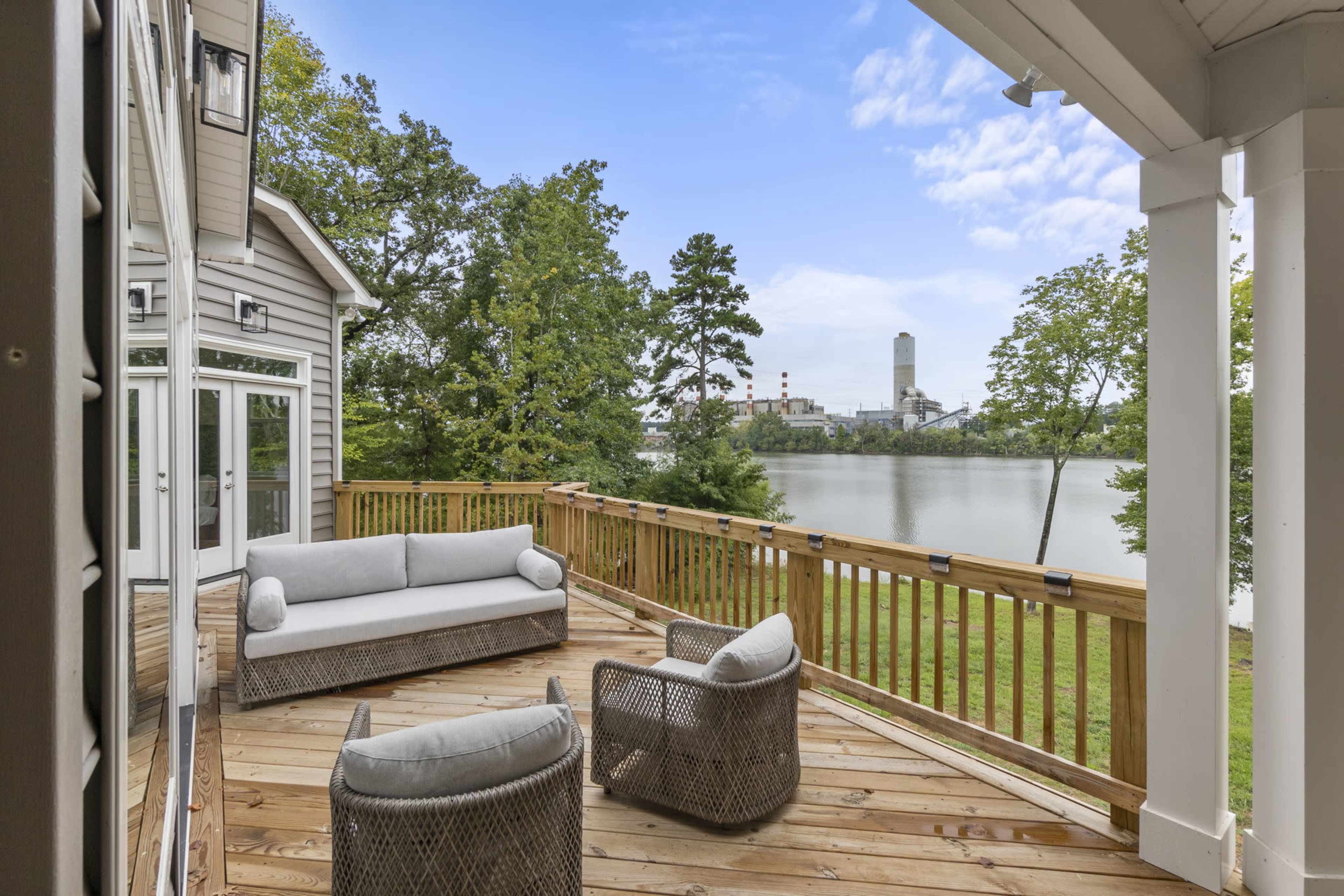 A deck with a sofa and two armchairs overlooks a body of water and industrial buildings in the distance.