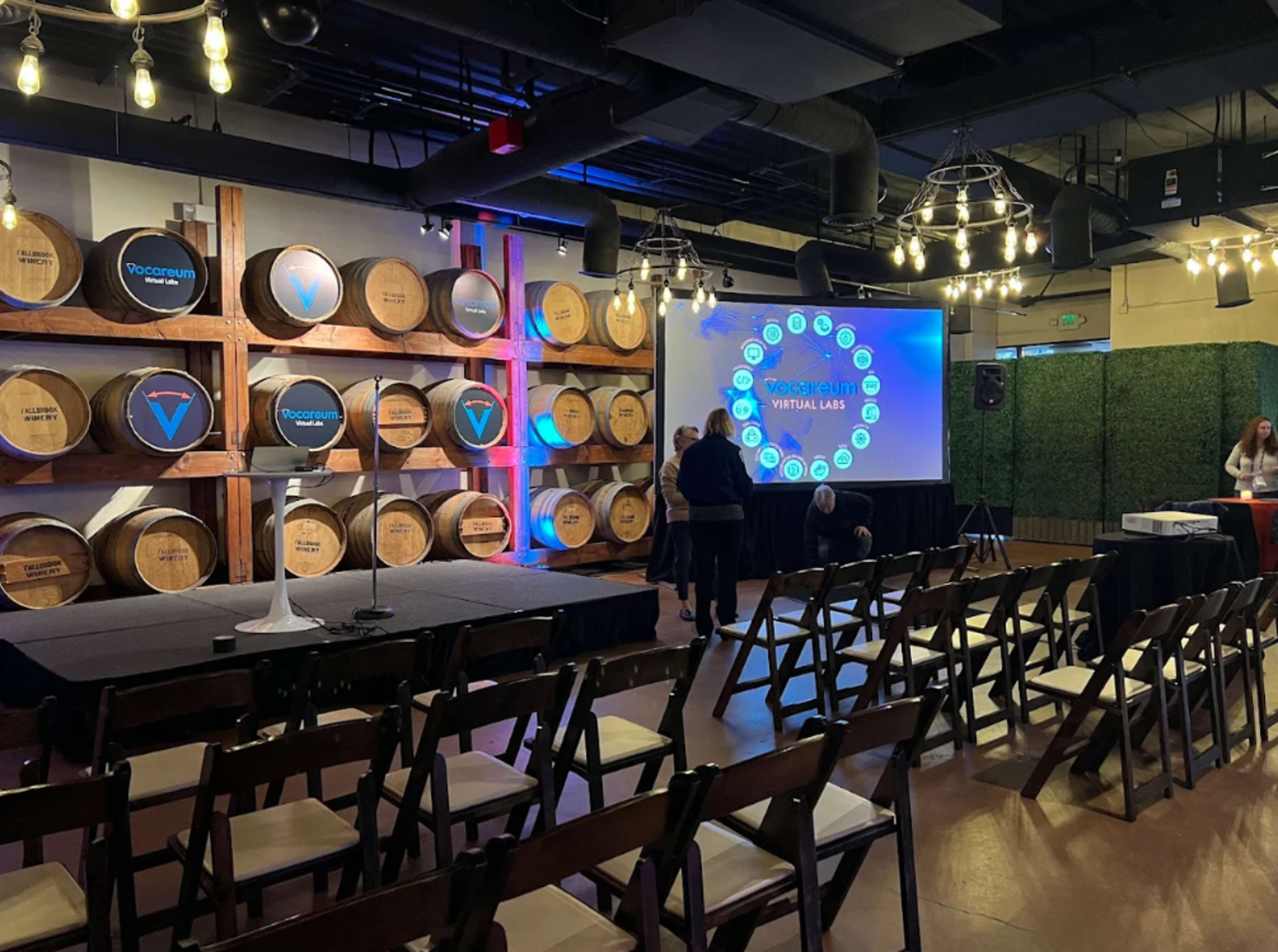 The image shows a preparation area for an event featuring a stage with a podium, surrounded by wooden barrels and illuminated by soft lighting.