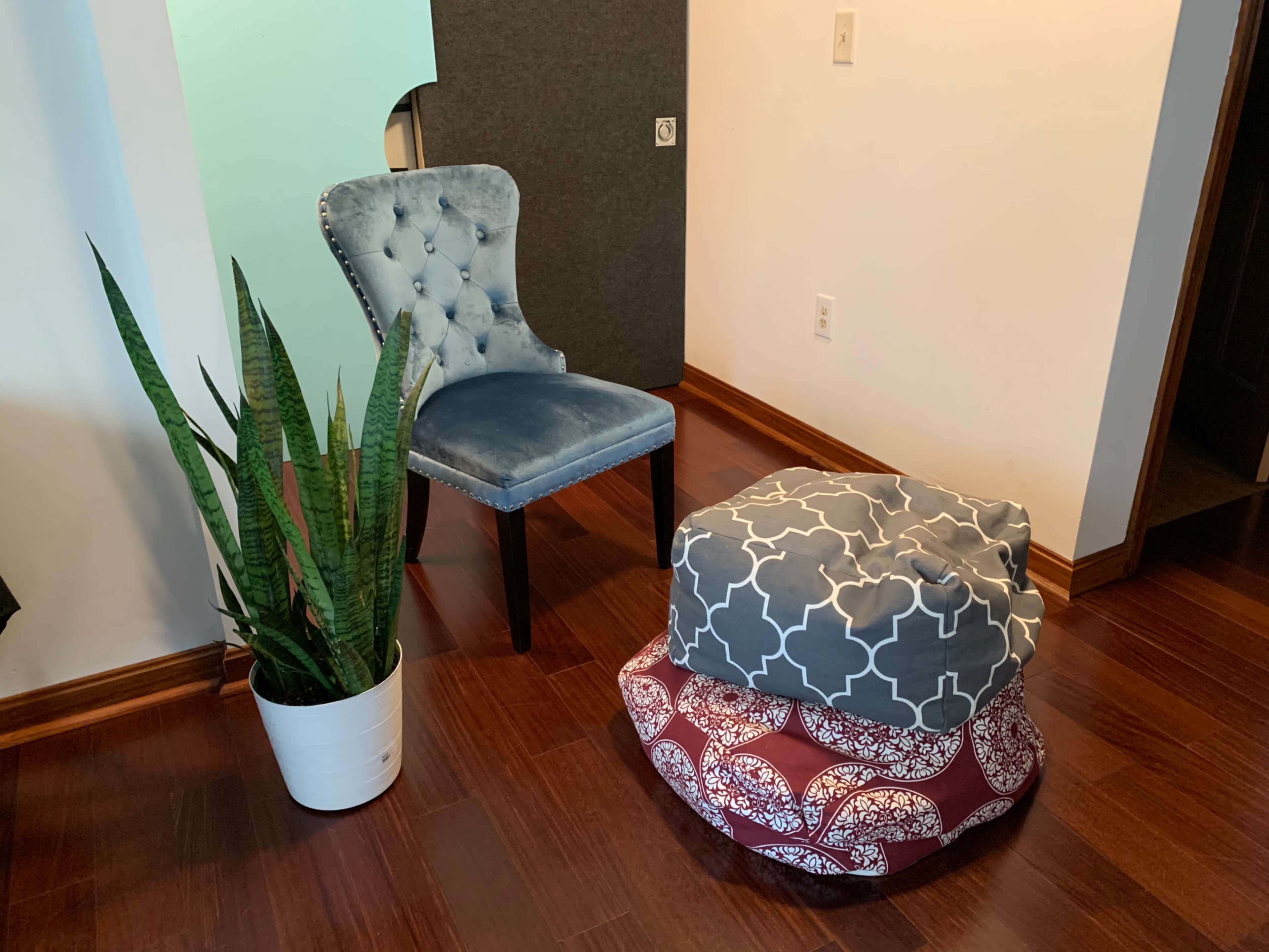 A blue upholstered chair and two patterned ottomans sit beside a tall potted plant in a room with hardwood floors.