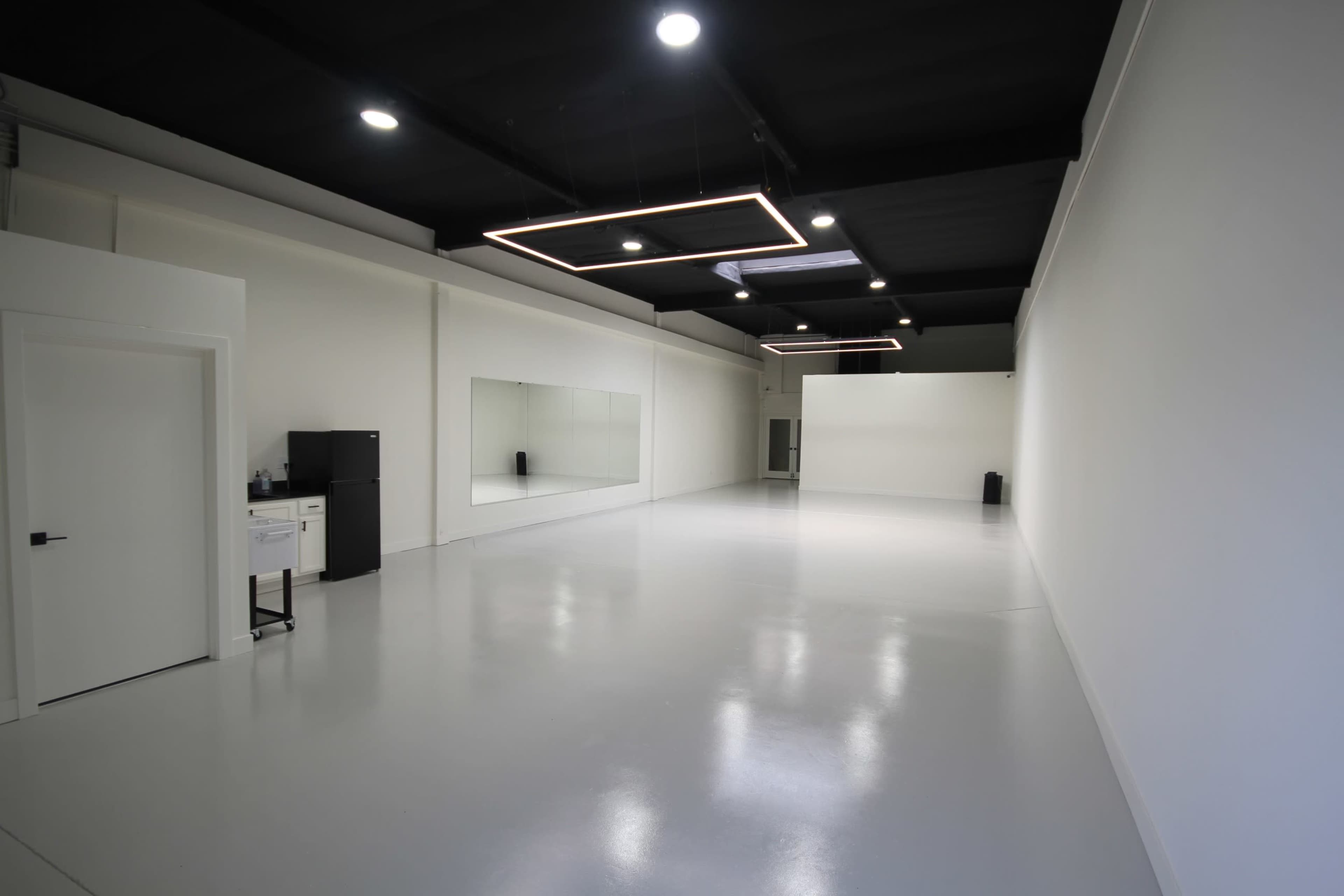 The image shows a spacious, empty room with white walls, a large mirror, and modern lighting fixtures.