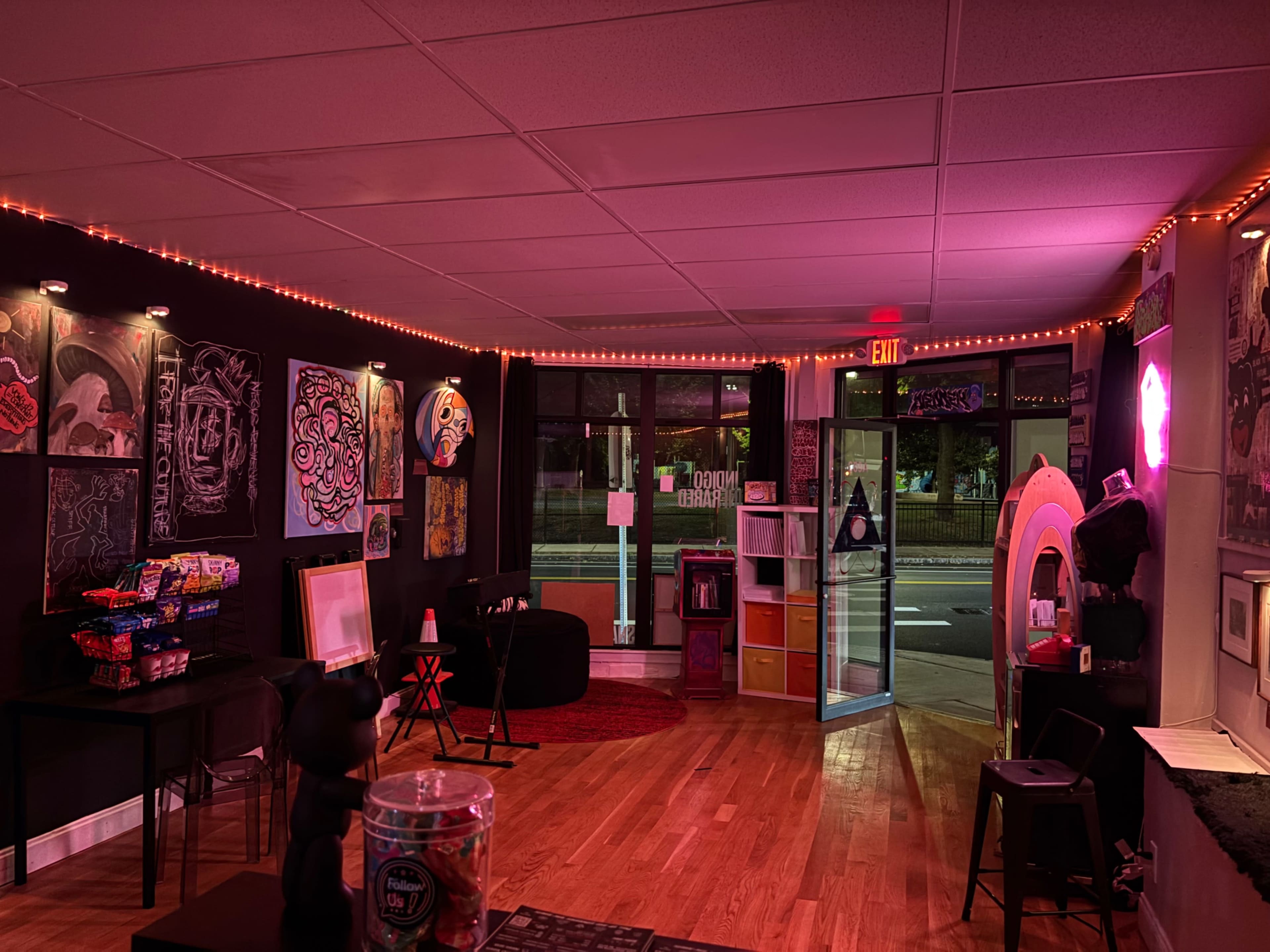 The image shows an art gallery or studio space with black walls adorned with colorful artwork, soft lighting, and various furniture pieces scattered throughout the room.