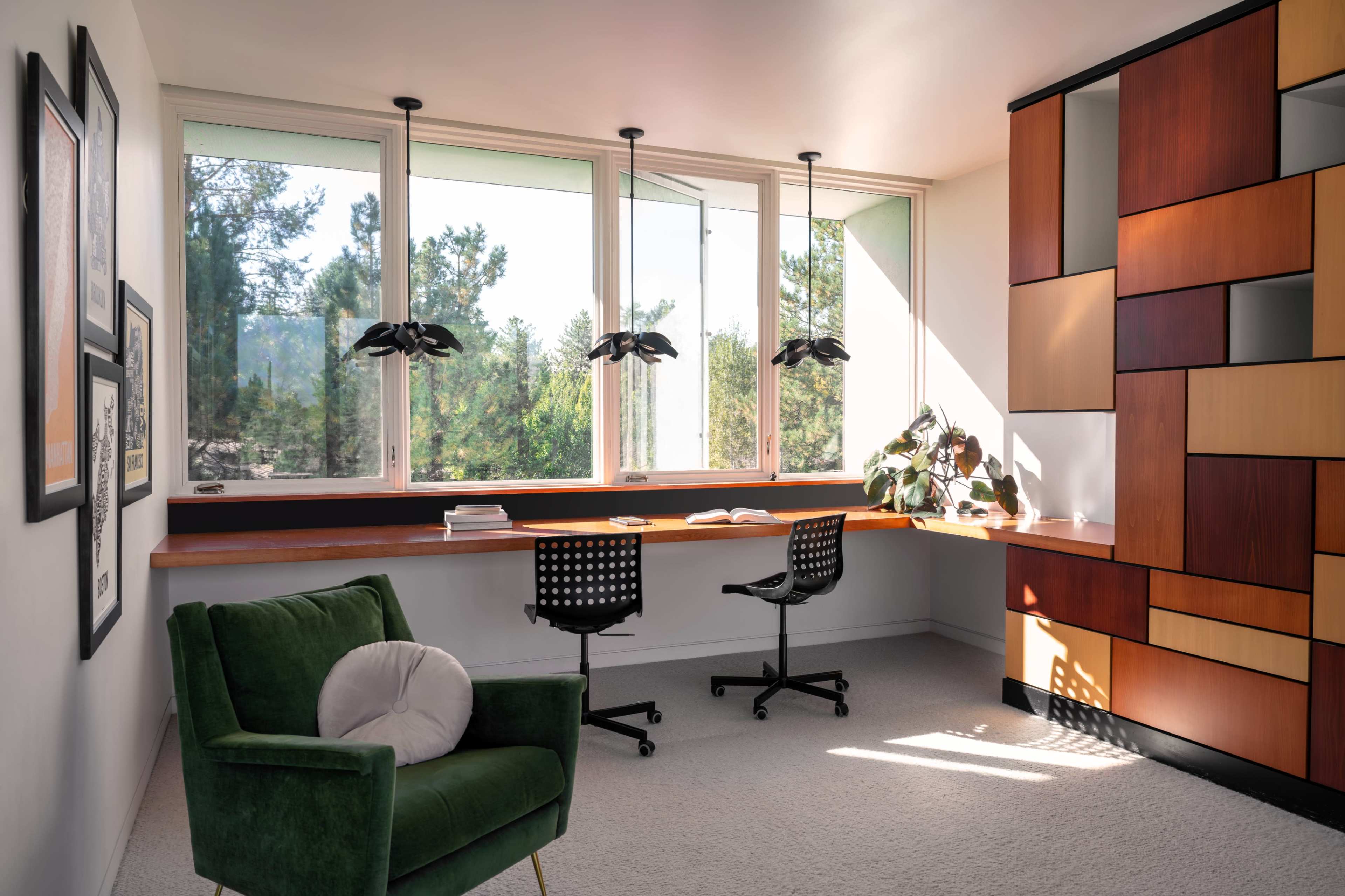 A modern home office features a long desk with two black chairs, large windows revealing trees outside, and a green armchair in the corner.