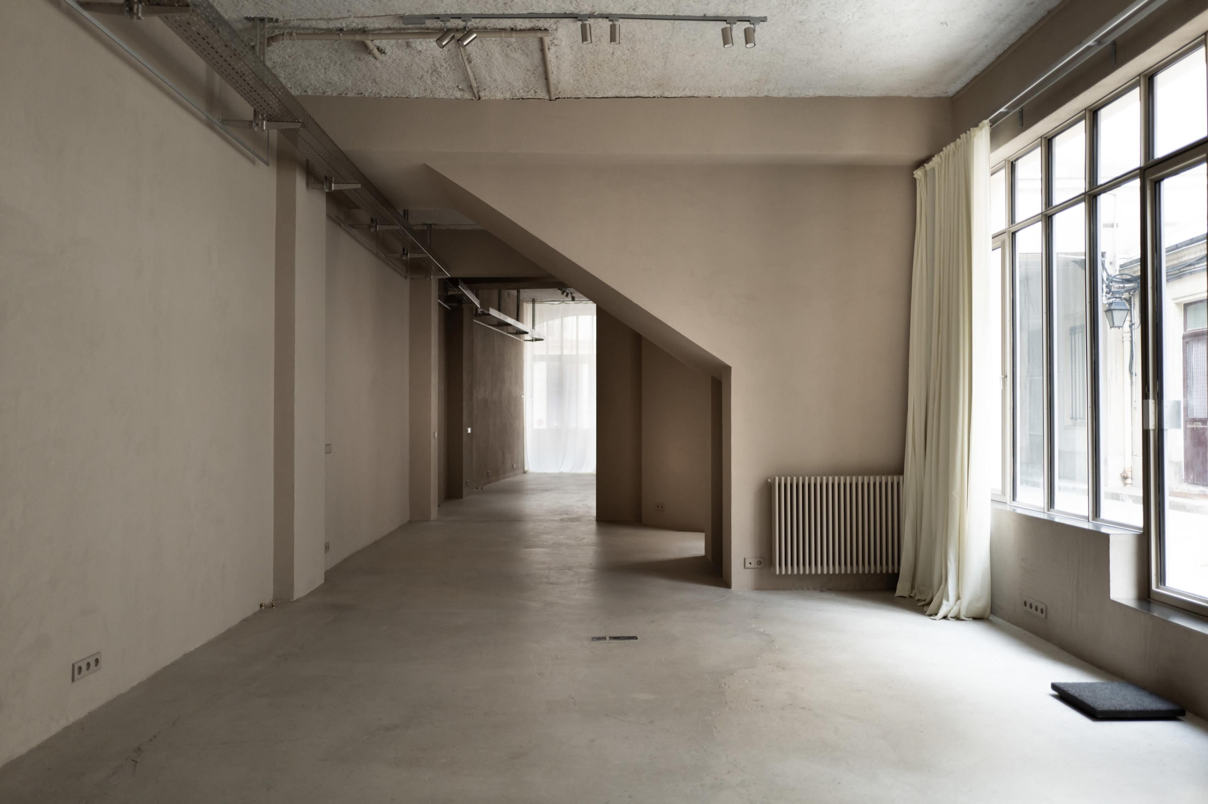 The image shows a spacious, empty room with soft gray walls, a large window, and an angular ceiling.
