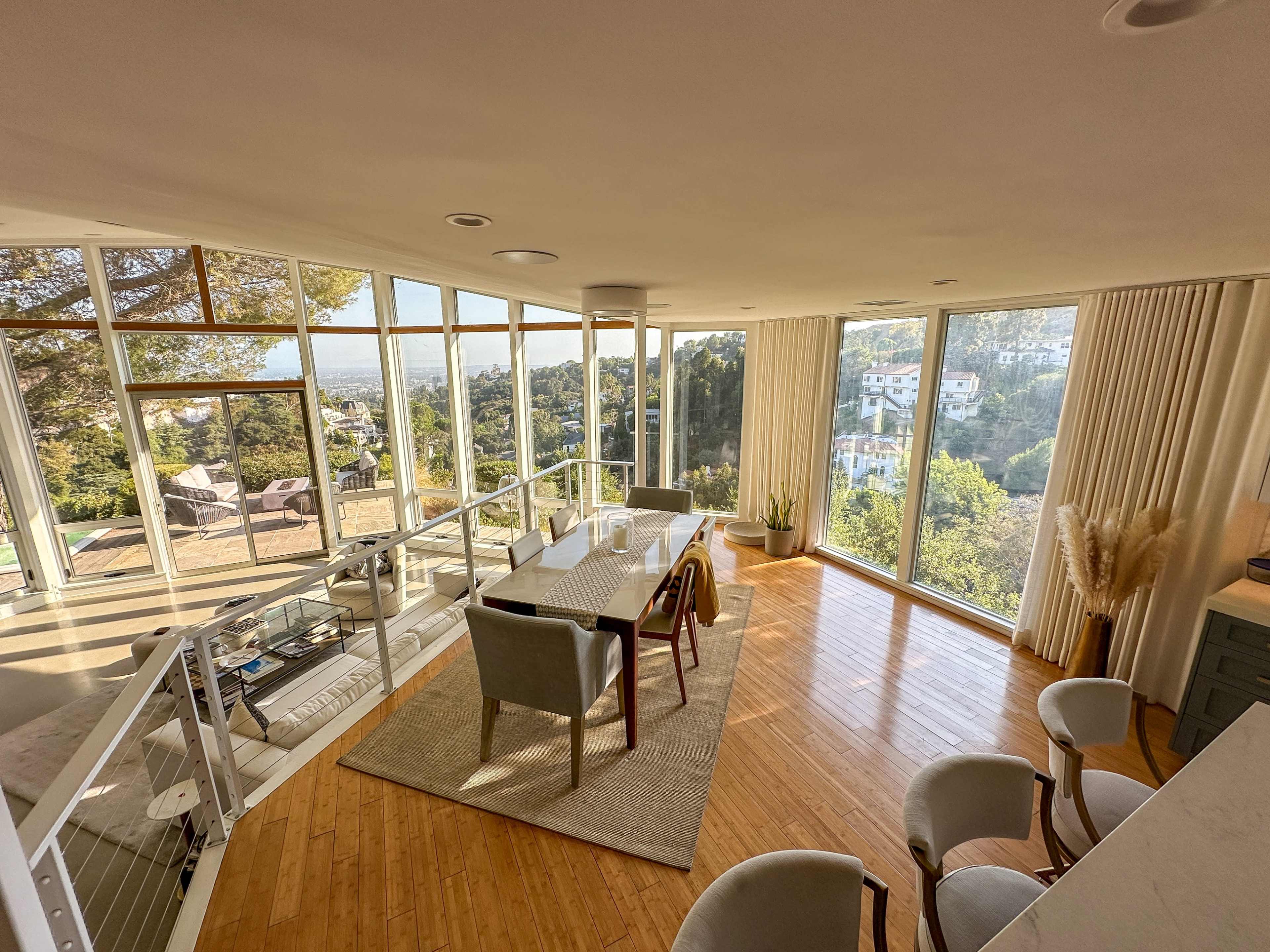 The image shows a modern dining area with large windows overlooking a hillside landscape, featuring a rectangular table and several chairs on a wooden floor.