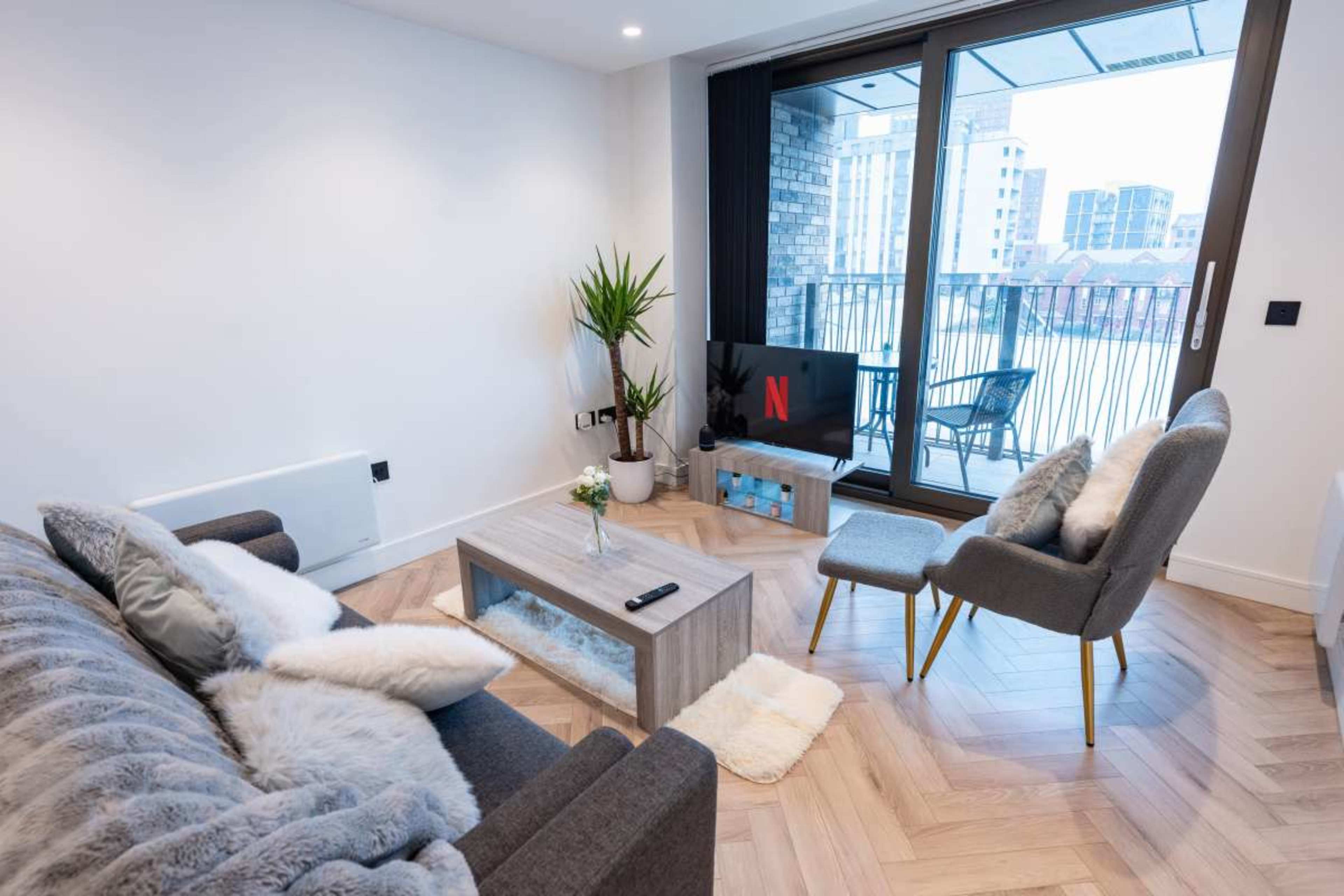 A modern living room features a gray sofa and armchair, a coffee table, and a television displaying the Netflix logo, with large windows revealing an urban view.
