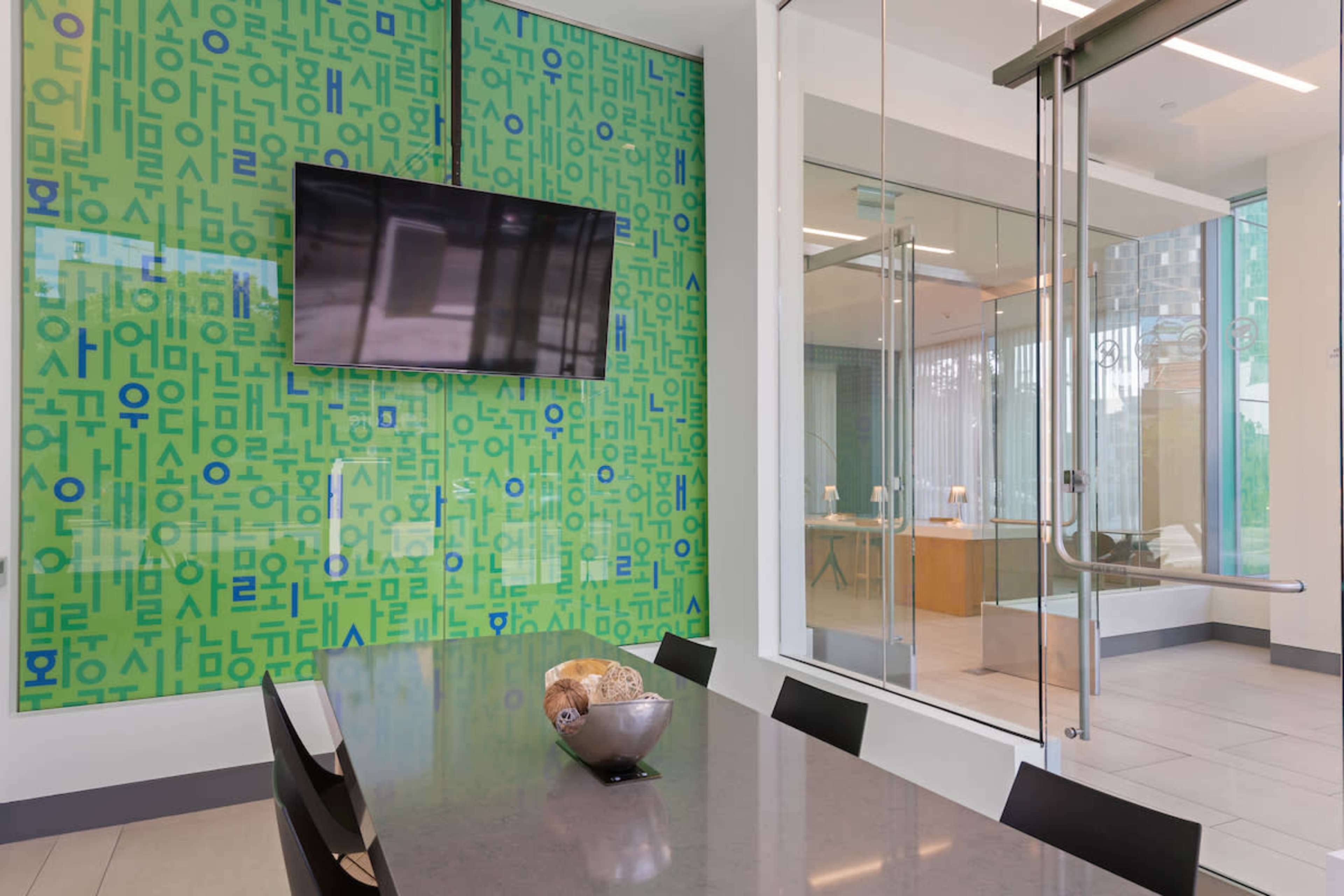 The image shows a modern conference room with a large table, a television mounted on a green wall covered in text, and glass doors leading to another room.