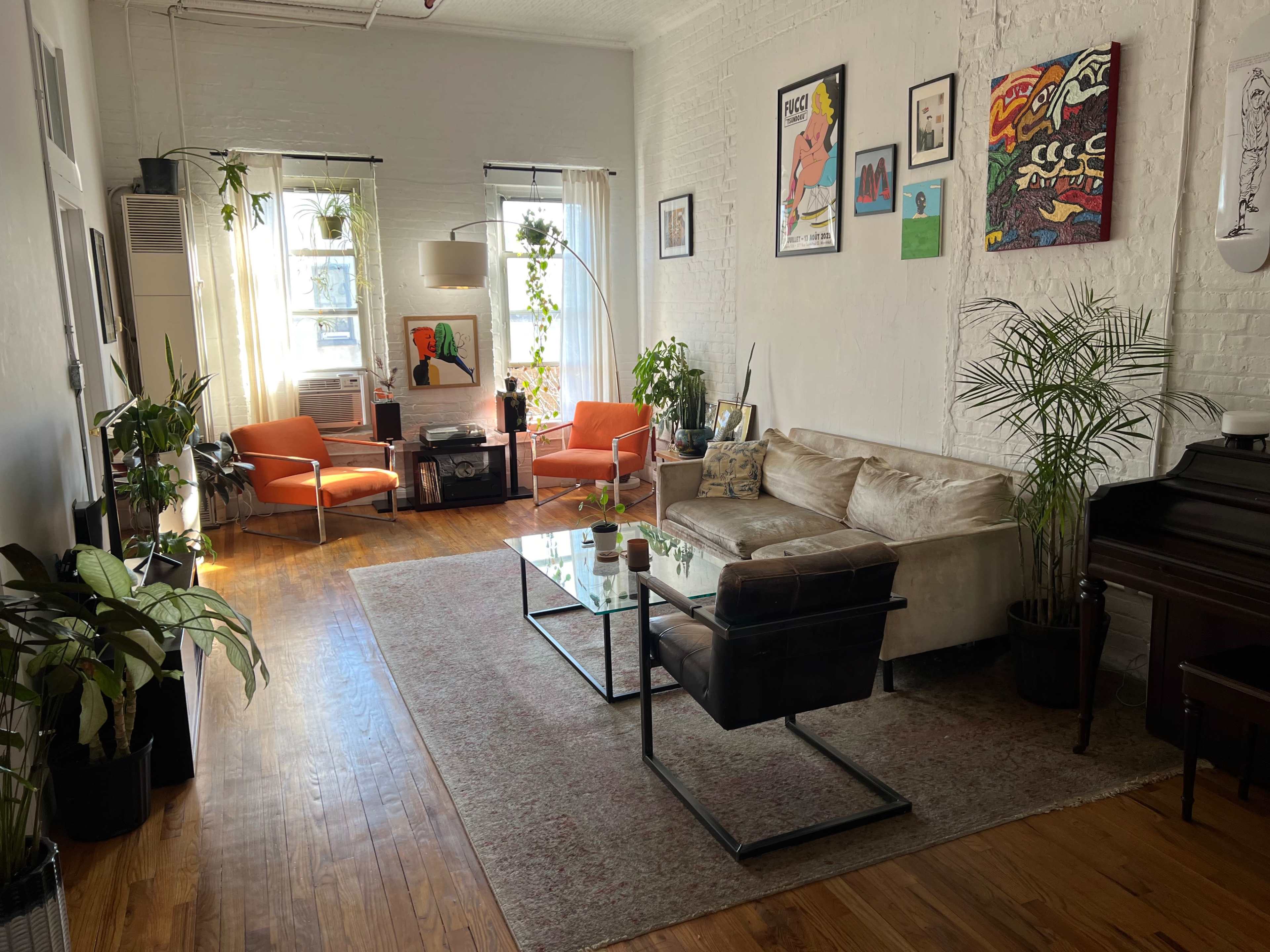 The image shows a bright living room with wooden floors, two orange chairs, a beige sofa, a coffee table, and various indoor plants, along with artwork on the walls.