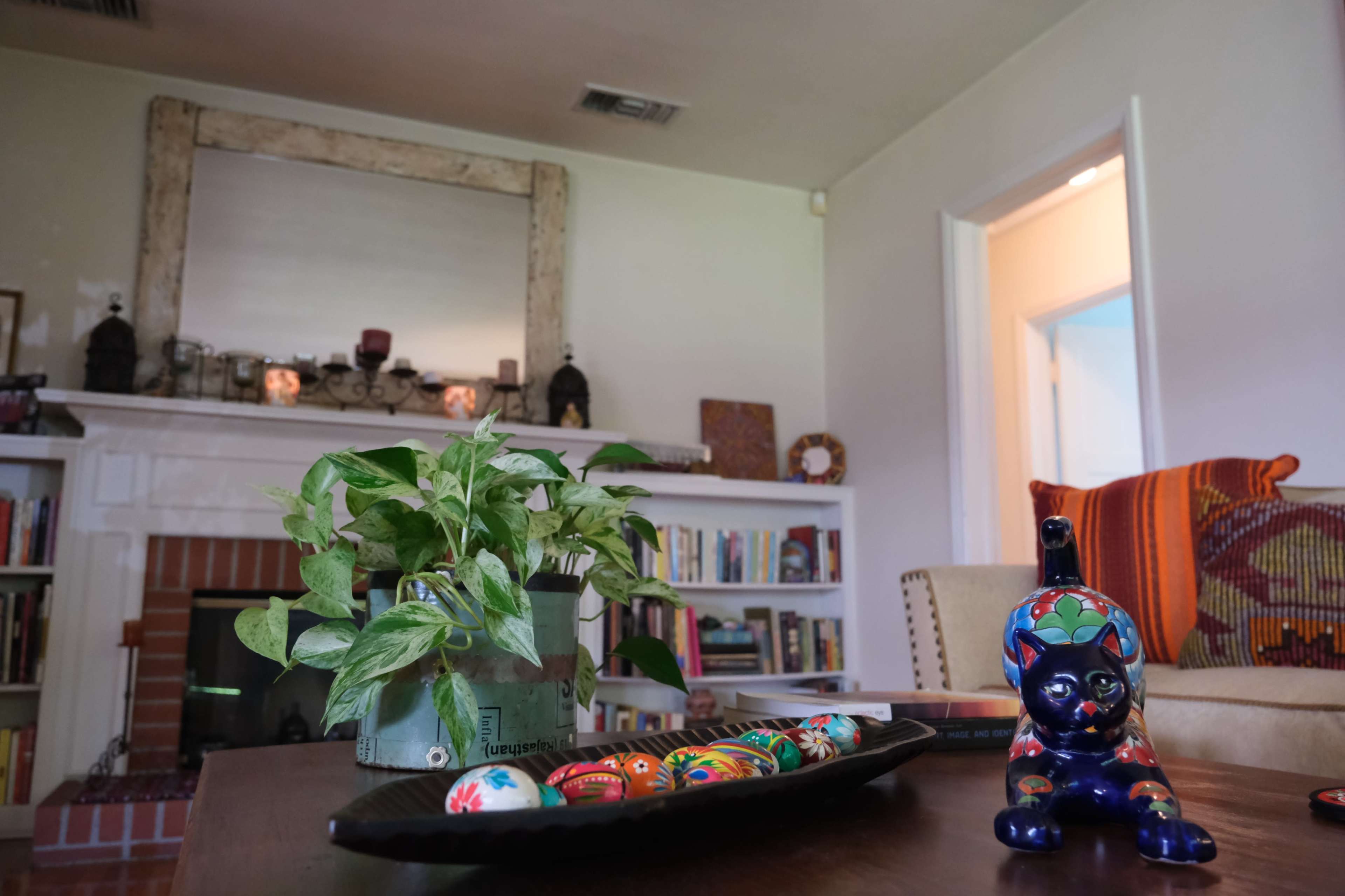 A living room features a decorative mantle with candles, a stack of books on a shelf, a potted plant on a table, and a colorful ceramic cat next to a tray of ornamental eggs.