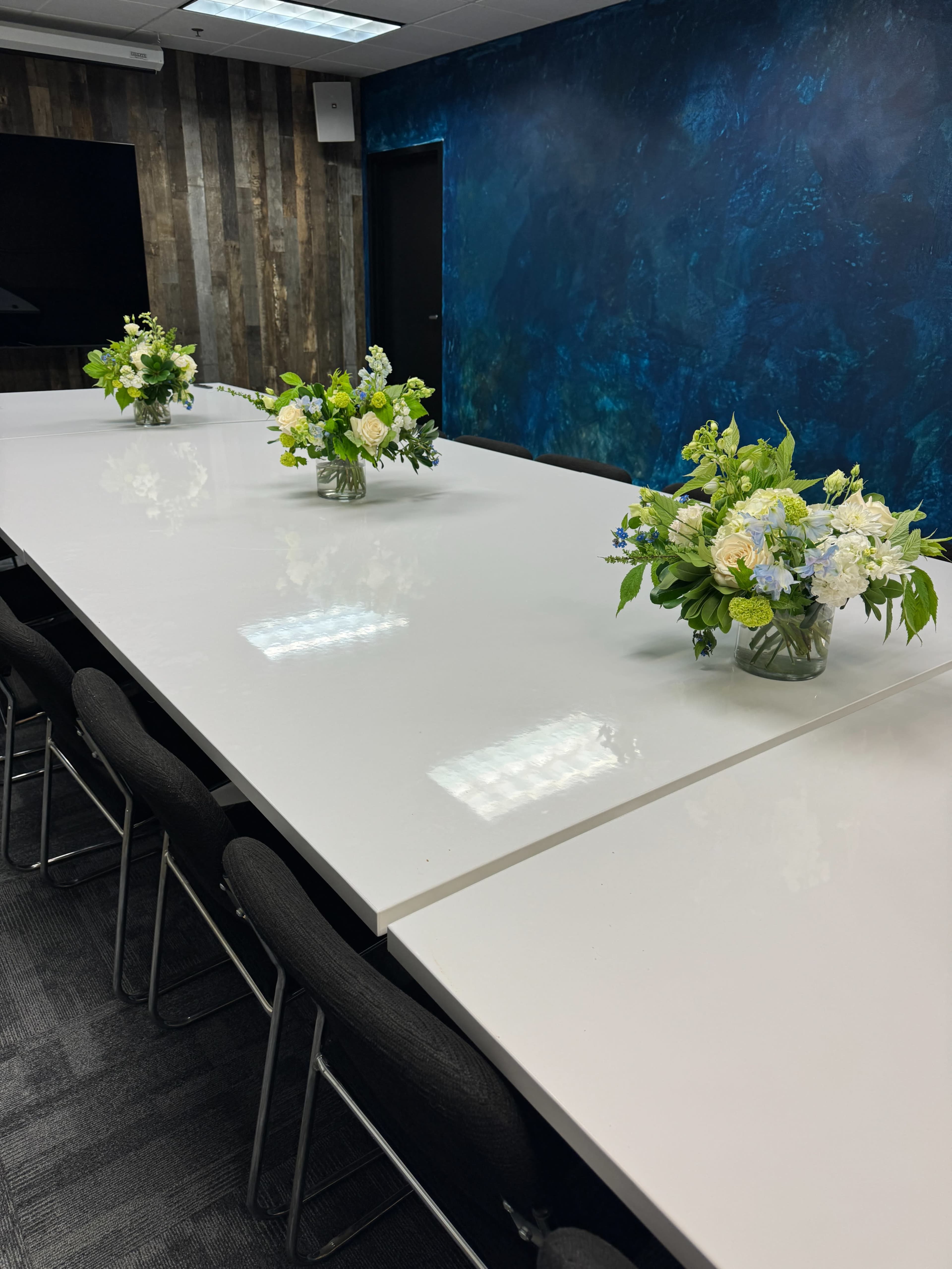 A conference room features a long white table adorned with flower arrangements at both ends and a dark blue textured wall.