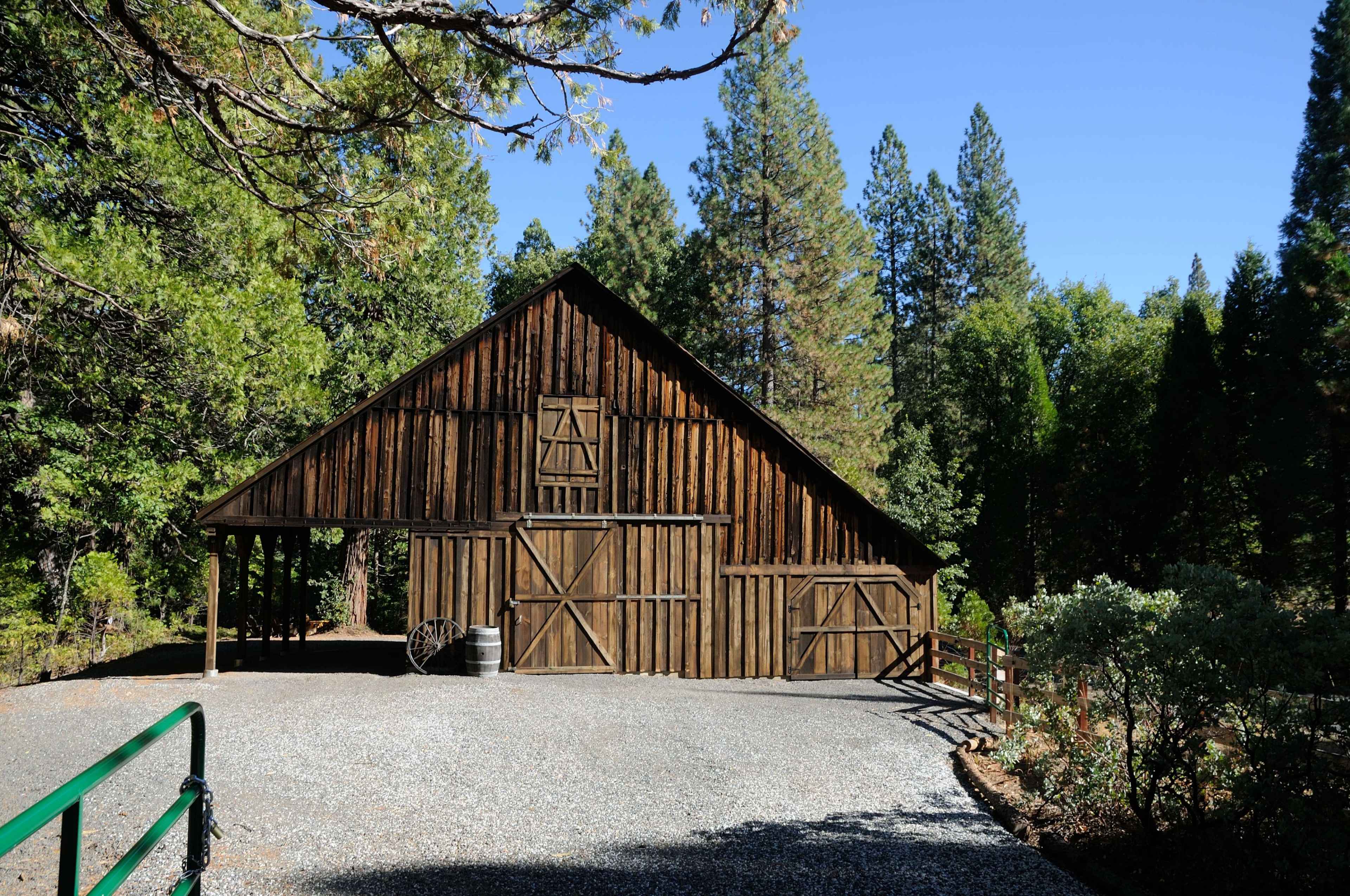 A wooden barn with a peaked roof stands next to a gravel driveway, surrounded by tall trees.