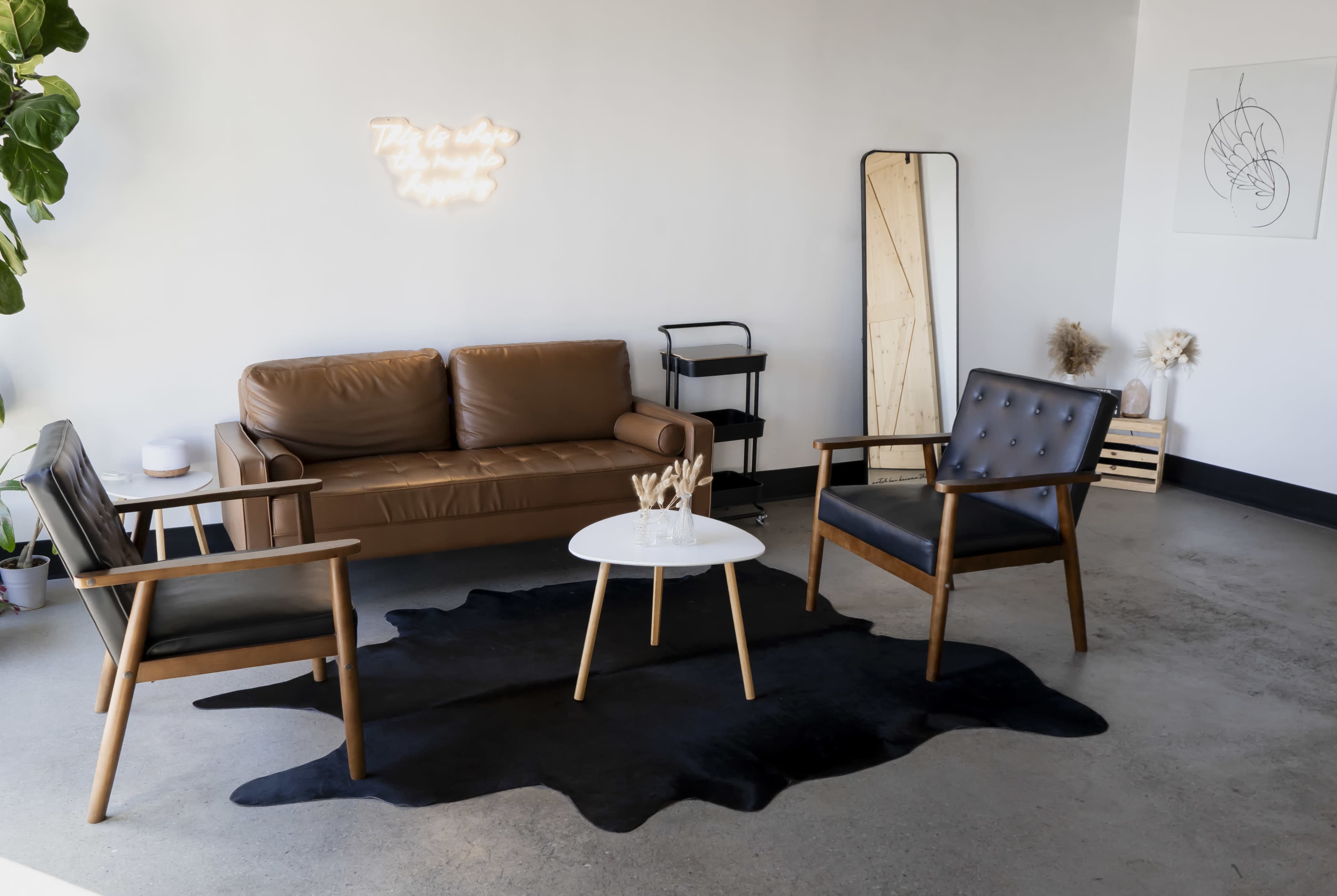 The image depicts a modern living space featuring a brown leather sofa, two black wooden chairs, a white round coffee table, and a large black area rug.