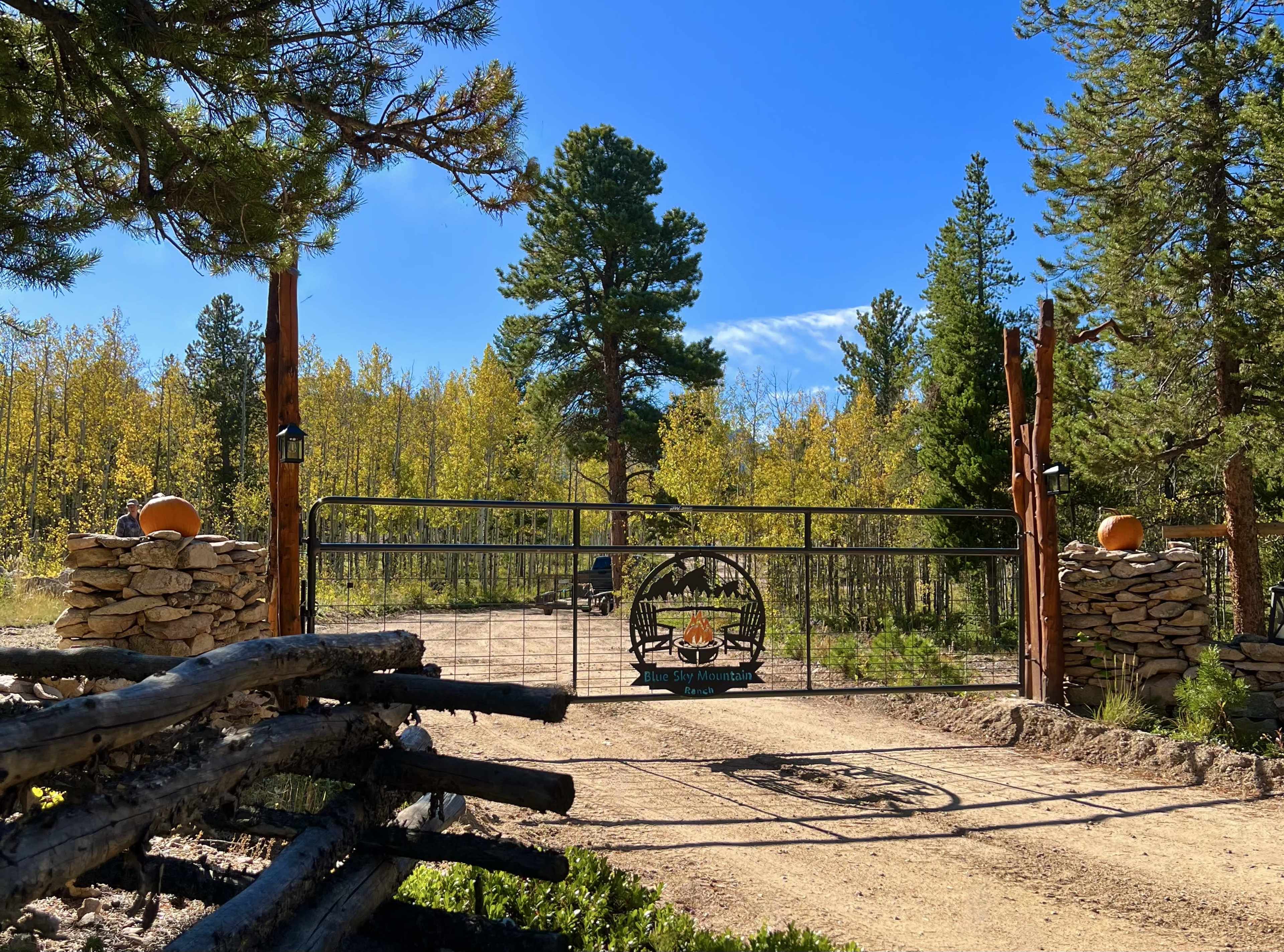 A metal gate with a decorative emblem stands at the entrance of a gravel road surrounded by tall trees and autumn-colored foliage.