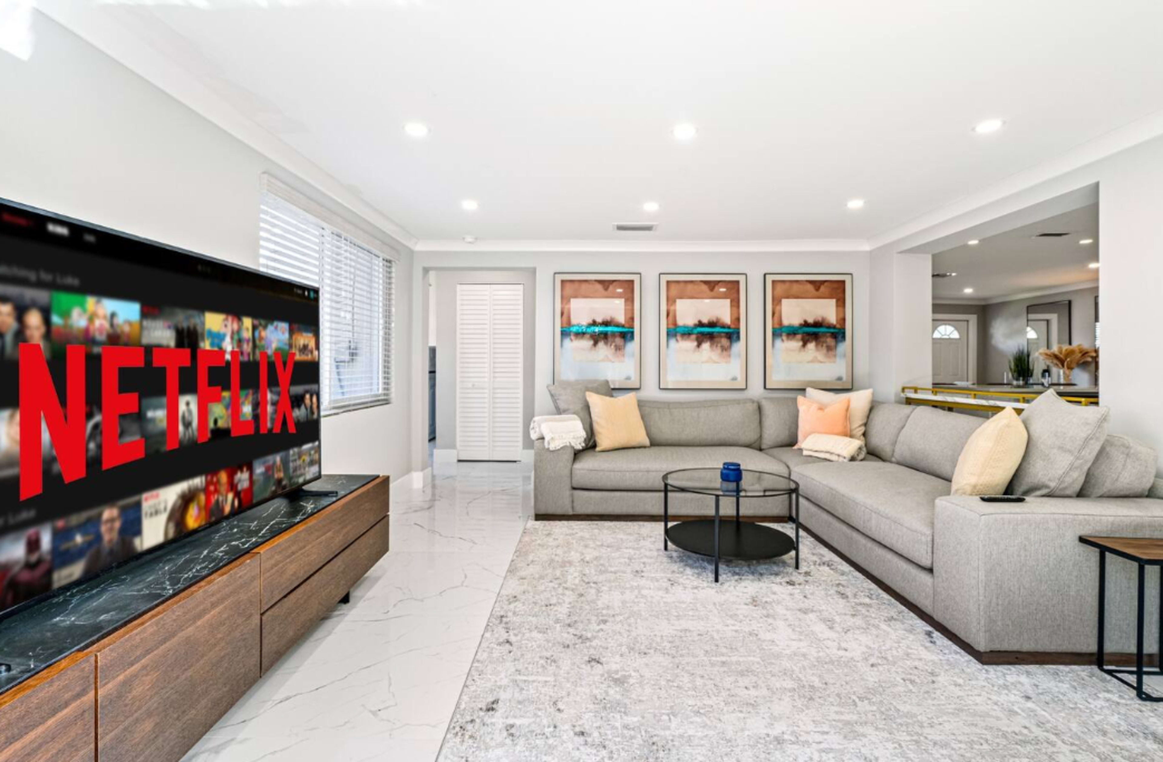 A modern living room features a gray sectional sofa, a coffee table, and a television displaying the Netflix logo, with light-colored walls and artwork on display.