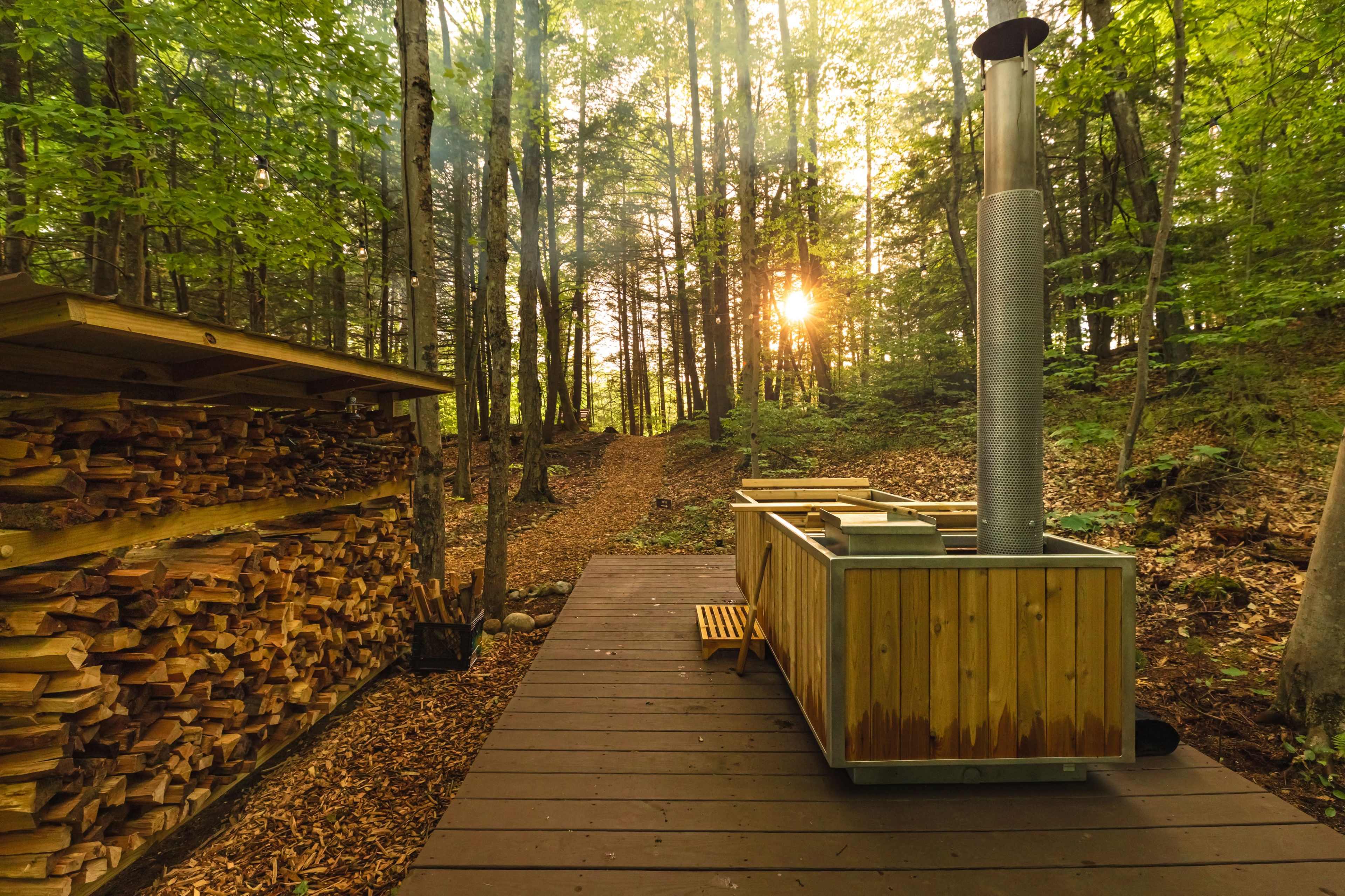 A wooden hot tub sits on a deck surrounded by trees, with a stack of firewood nearby and sunlight streaming through the foliage.
