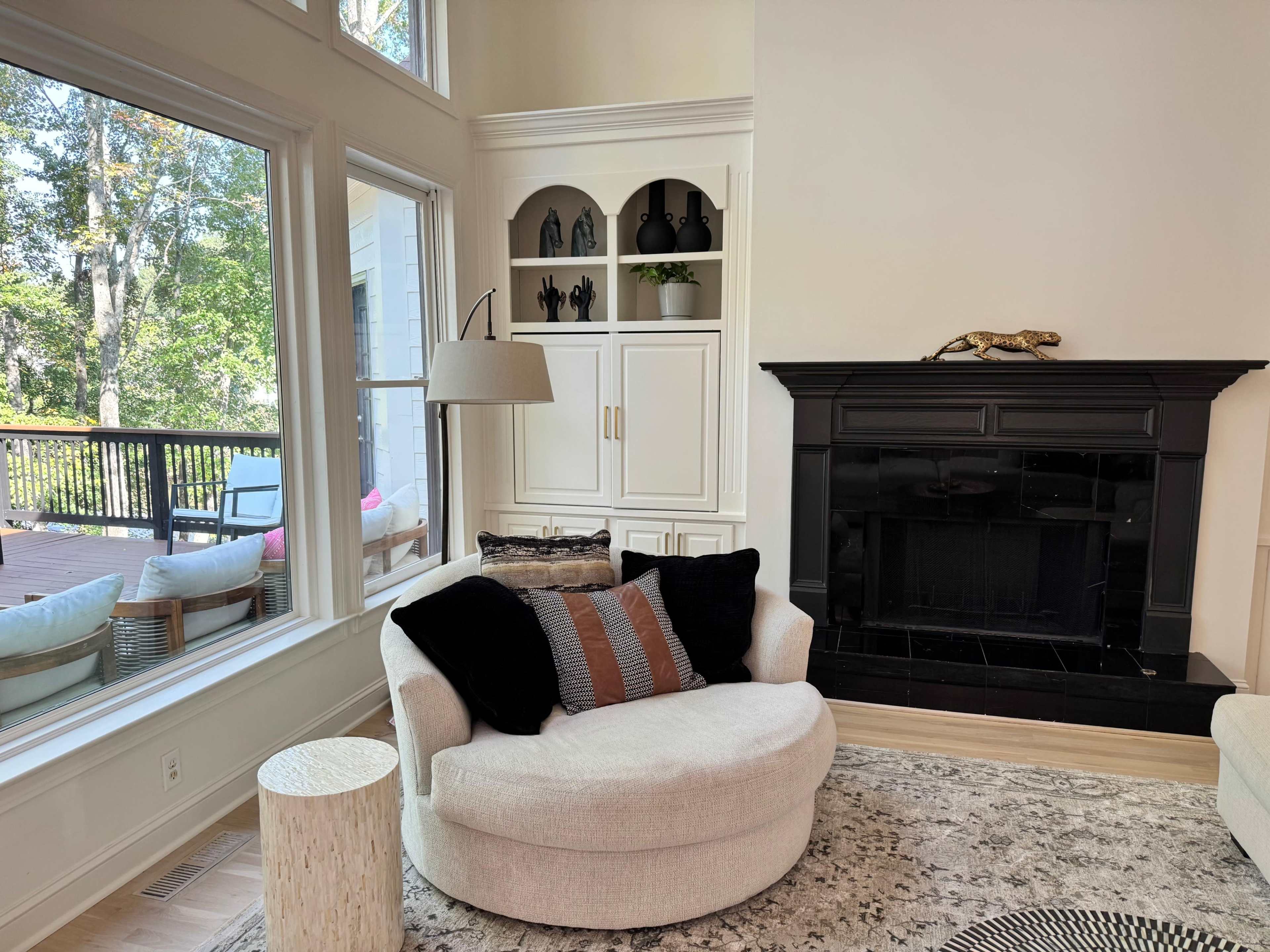 The image shows a cozy living room featuring a white upholstered chair, a black fireplace, and large windows overlooking a deck with outdoor seating.