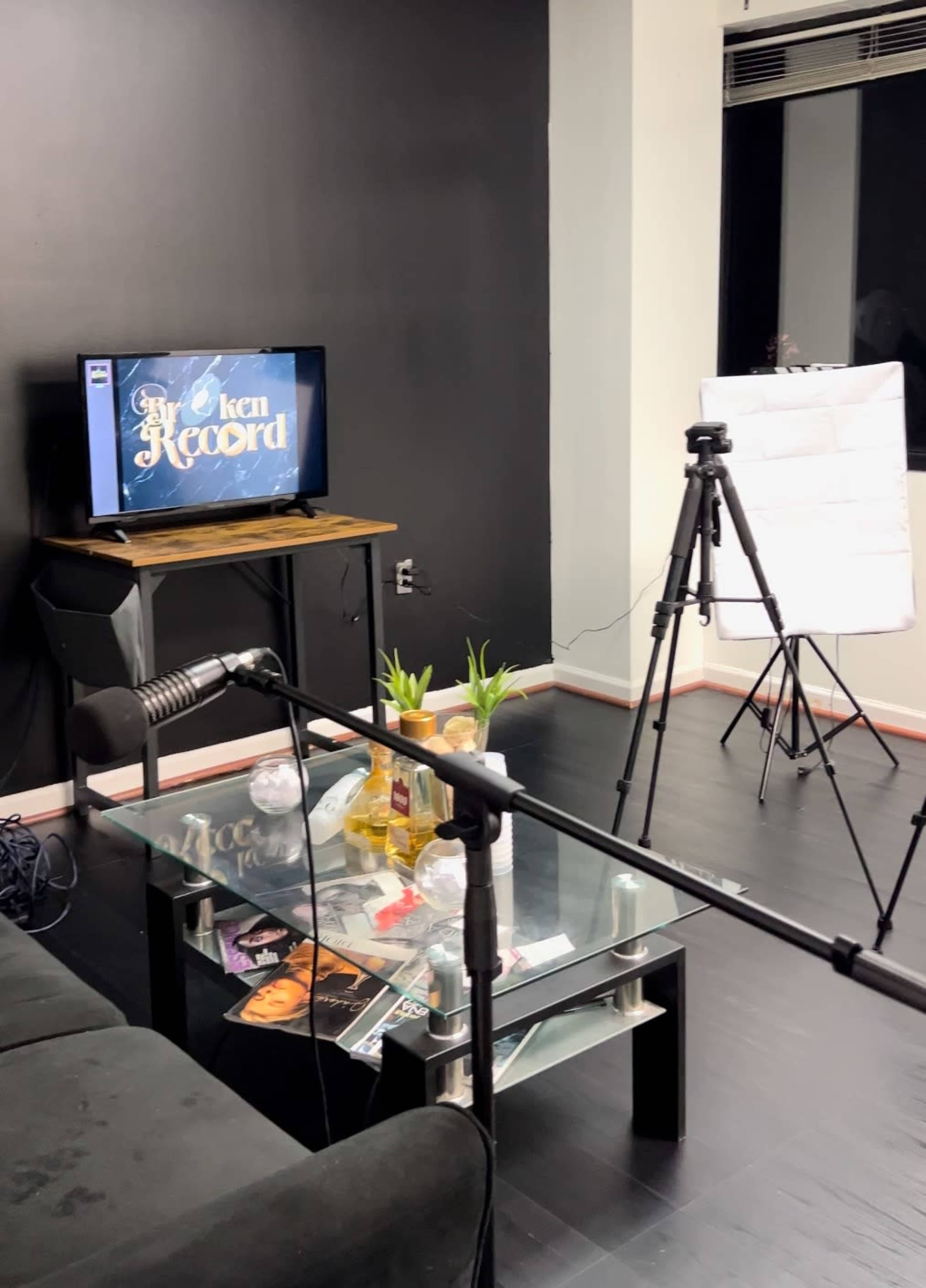 A modern recording studio setup features a black wall, a television displaying "Broken Record," a glass coffee table with plants and items, and equipment for filming.