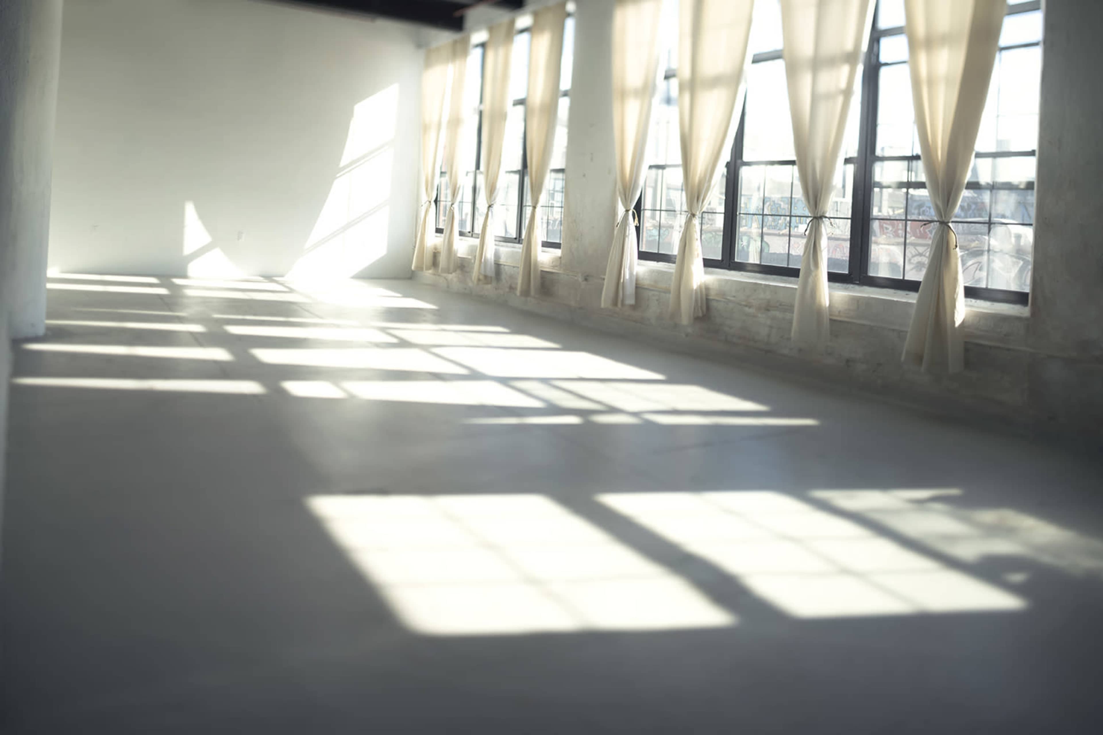 The image shows a spacious room with large windows draped in light curtains, casting shadows on the polished floor.