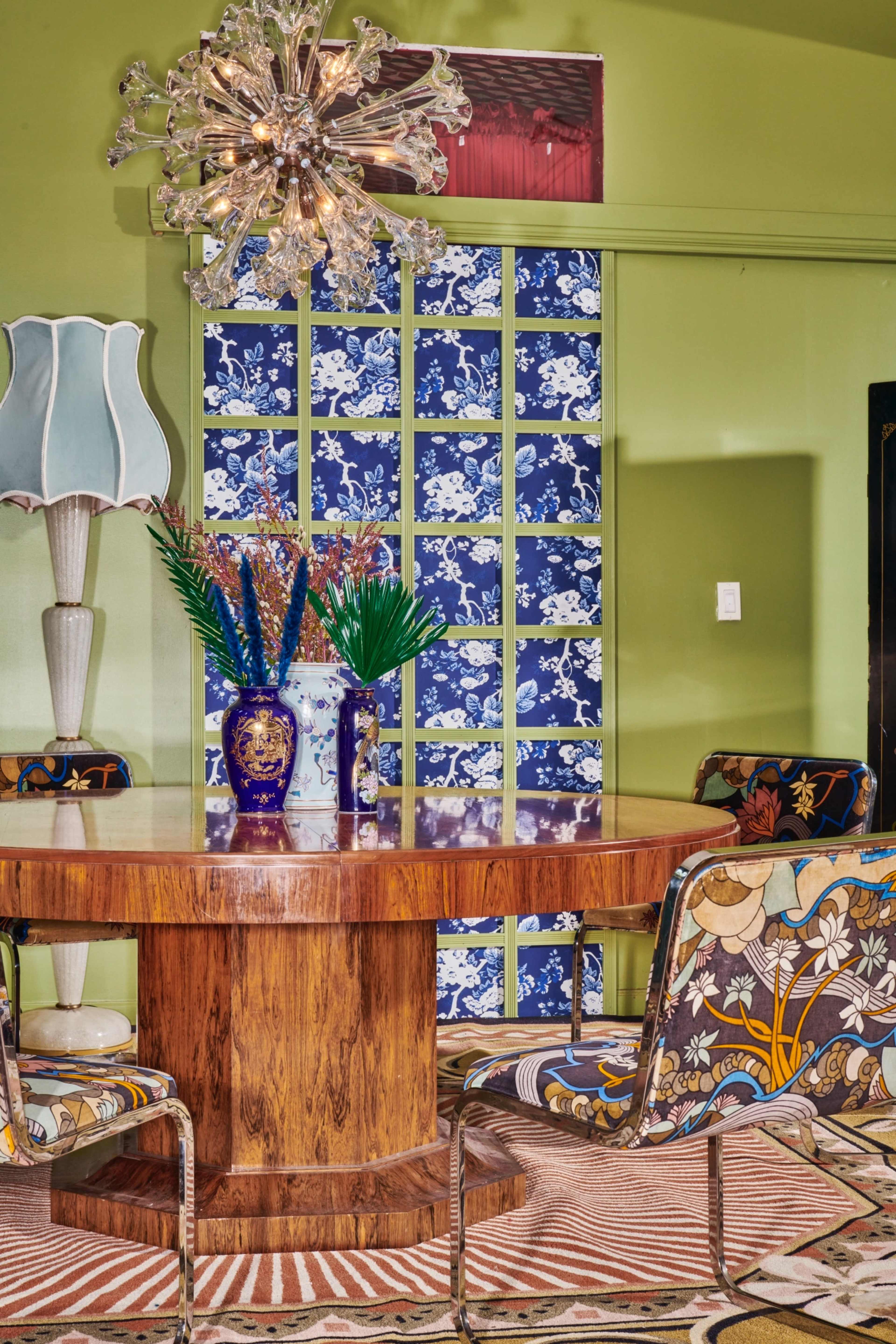 A round wooden dining table is surrounded by four patterned chairs, with a decorative lamp and a floral vase as centerpieces against a backdrop of blue and white wallpaper.