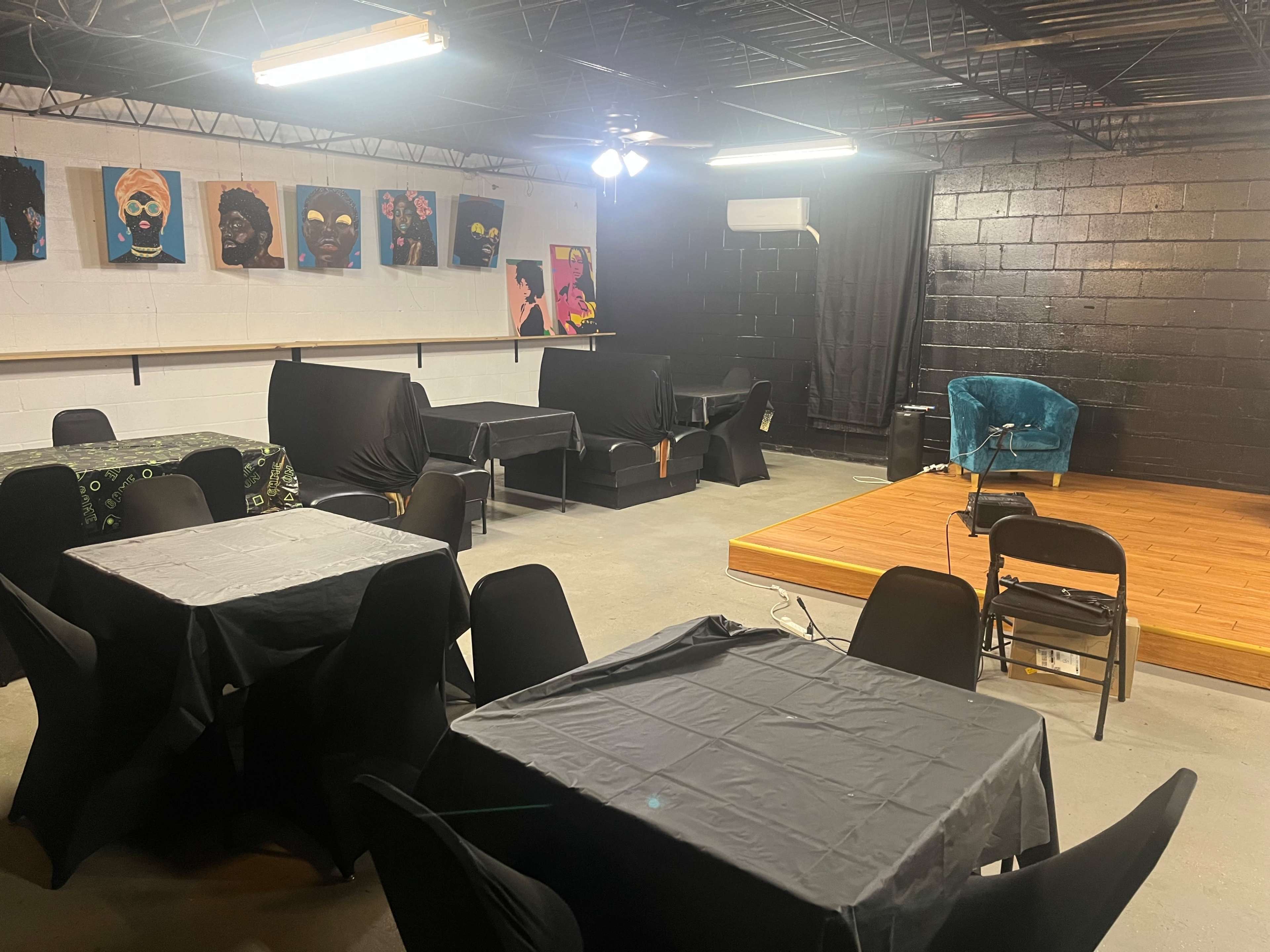 A dimly lit multi-purpose room with black tablecloths on several tables, a small stage area, and colorful wall art featuring portraits.