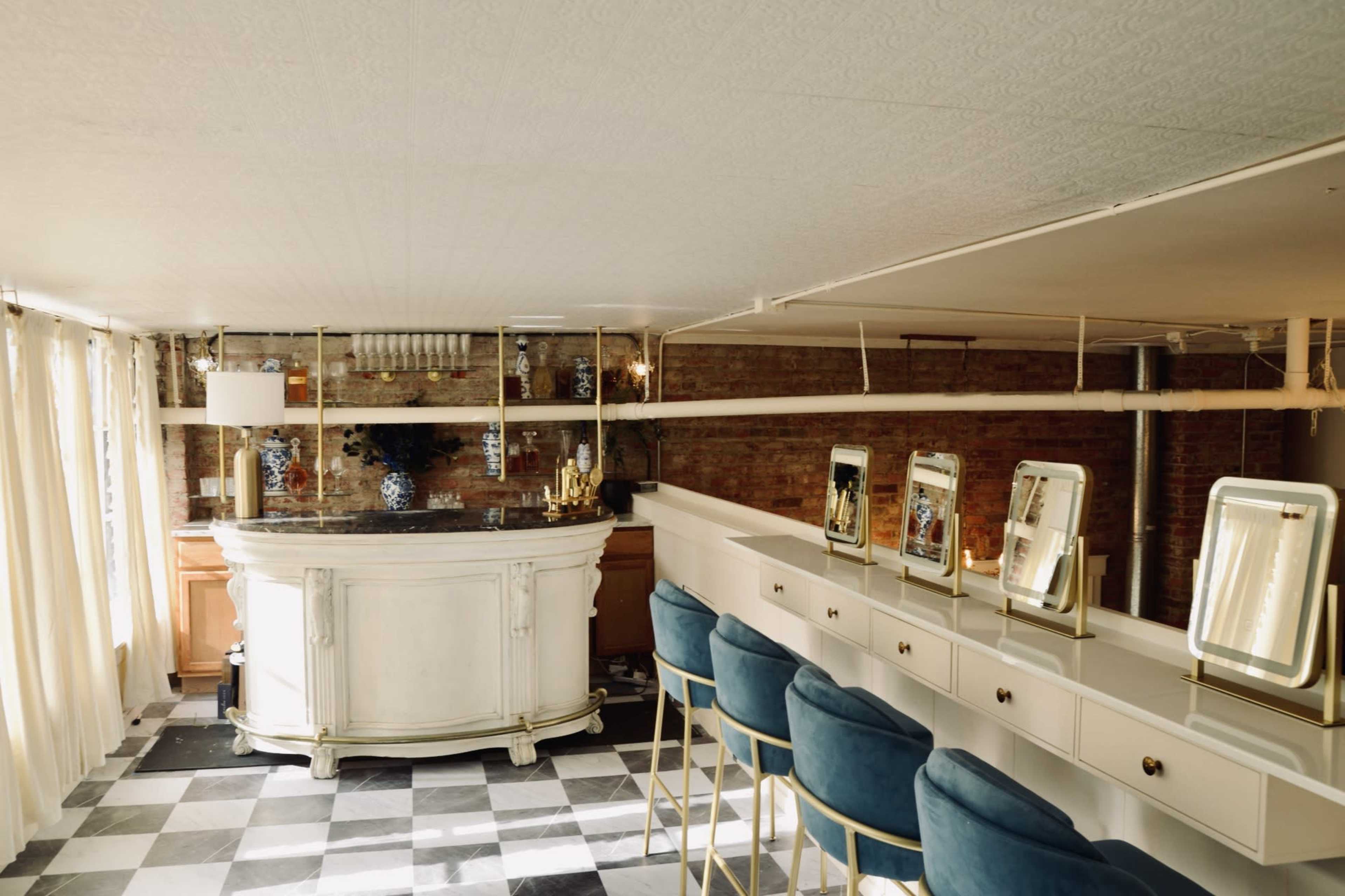 A bright interior features a sleek bar area with a polished countertop and several illuminated mirrors along a white countertop, complemented by blue bar stools.