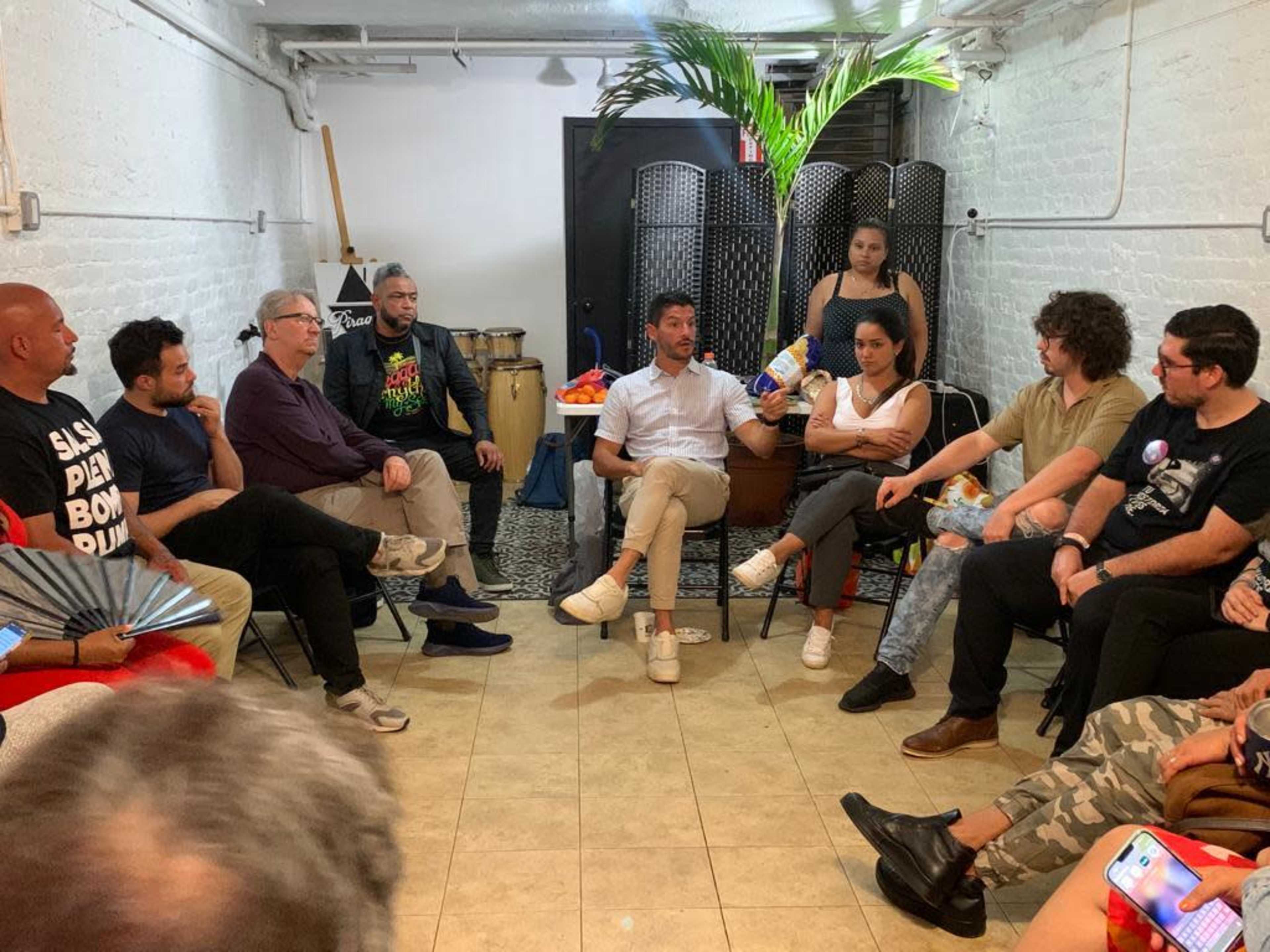 A group of people is seated in a circle in a bright room with plants, discussing while some musicians' instruments and fruit are visible in the background.